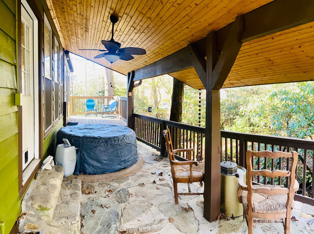A covered porch features a hot tub surrounded by stone paving and wooden railings. Two wooden chairs are positioned nearby, providing a comfortable seating area. Ceiling fans enhance the cooling effect, while trees and greenery are visible in the background.