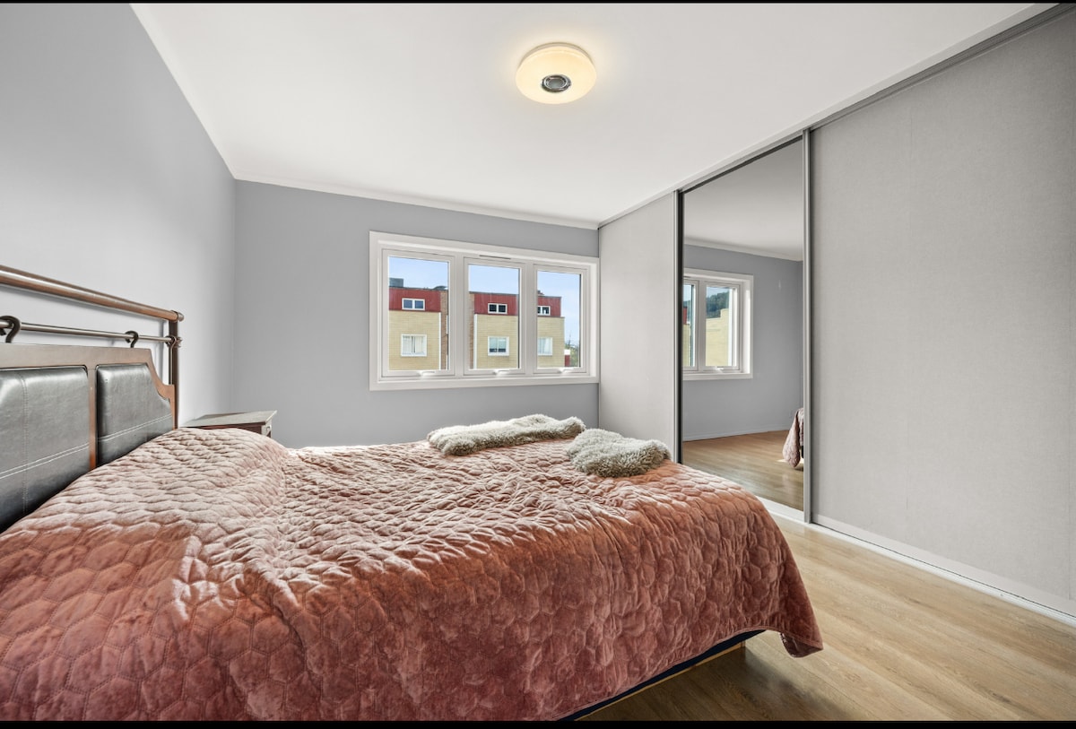 A spacious bedroom featuring a comfortable bed with a textured quilt and two soft throws. A large window allows natural light to fill the room, and a mirrored closet reflects the ample space. Soft grey walls create a neutral backdrop.
