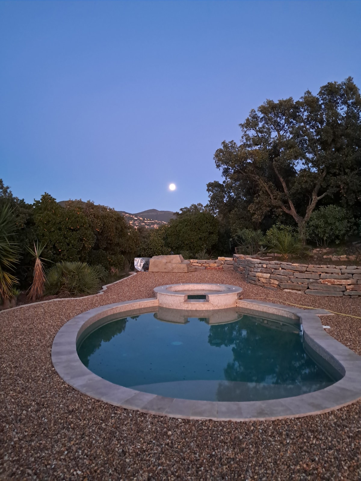 A neatly designed circular pool is surrounded by natural stone walls and gravel. Lush greenery is visible in the background, with trees creating a serene setting. The surface of the water reflects a soft moonlight, enhancing the tranquil ambiance of the outdoor area.