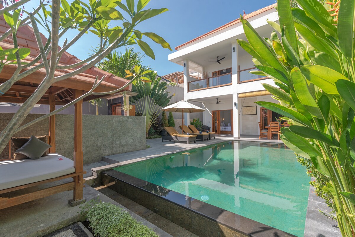 The image displays a serene outdoor space featuring an infinity pool surrounded by lush greenery and a daybed shaded by an umbrella. Two loungers are positioned by the pool, and a traditional gazebo is visible in the corner, adding to the tranquil ambiance.