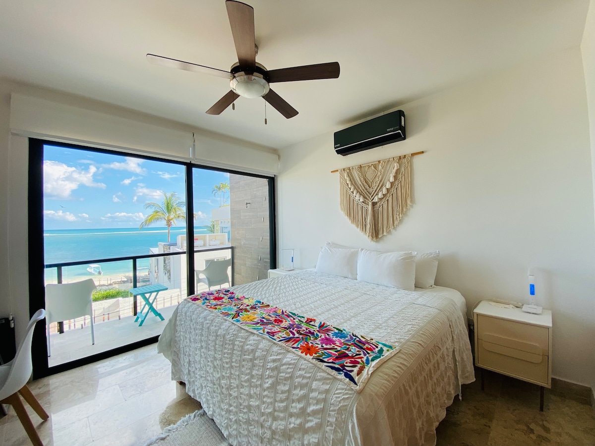 A spacious bedroom features a large bed with crisp white linens, accented by a colorful tapestry along the top. A ceiling fan and air conditioning unit are visible. Floor-to-ceiling glass doors provide a view of the ocean and lead to a balcony with outdoor seating.