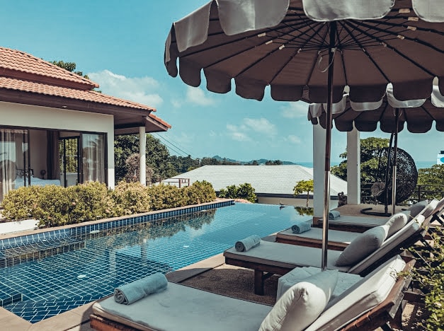 A serene pool area is displayed with four loungers arranged neatly beneath large umbrellas. The clear water of the pool reflects the blue sky, while landscaped greenery frames the space. A distant view of the sea can be seen through the surrounding trees.