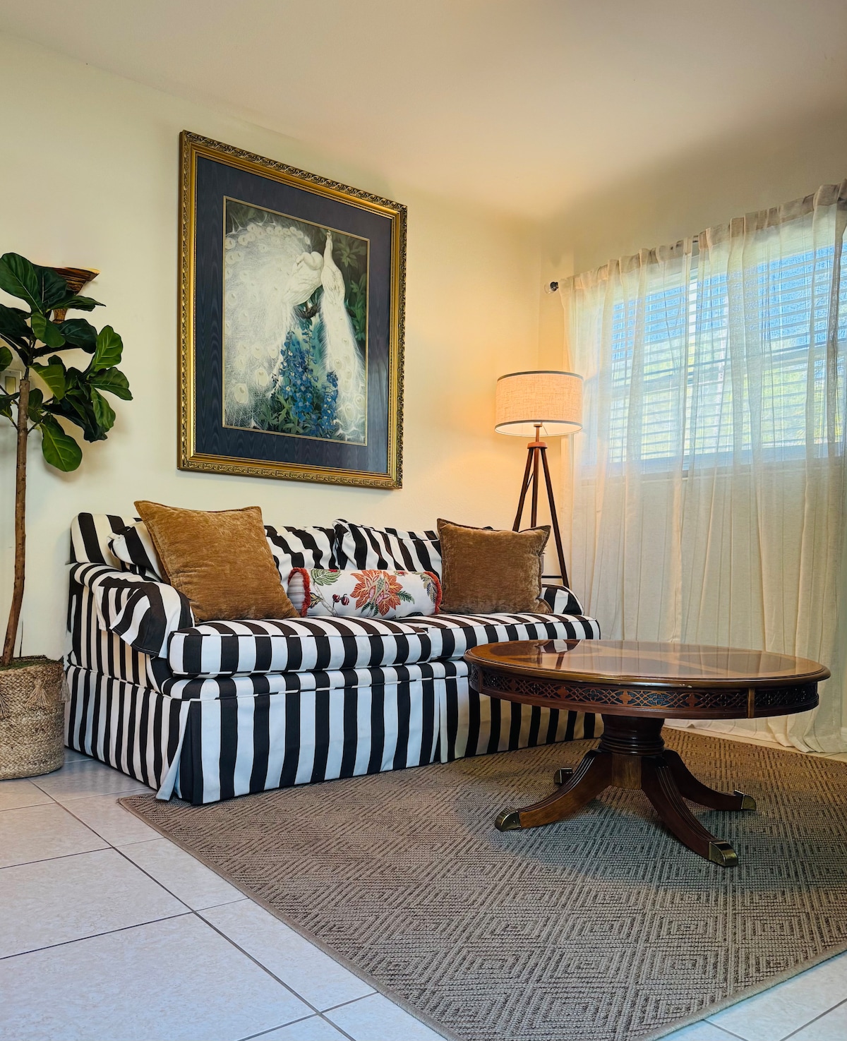 A striped sofa is positioned against a neutral wall, complemented by decorative cushions. A round wooden coffee table rests on a textured area rug. Natural light filters through sheer curtains, while a floor lamp adds soft illumination. A potted plant provides a touch of greenery.