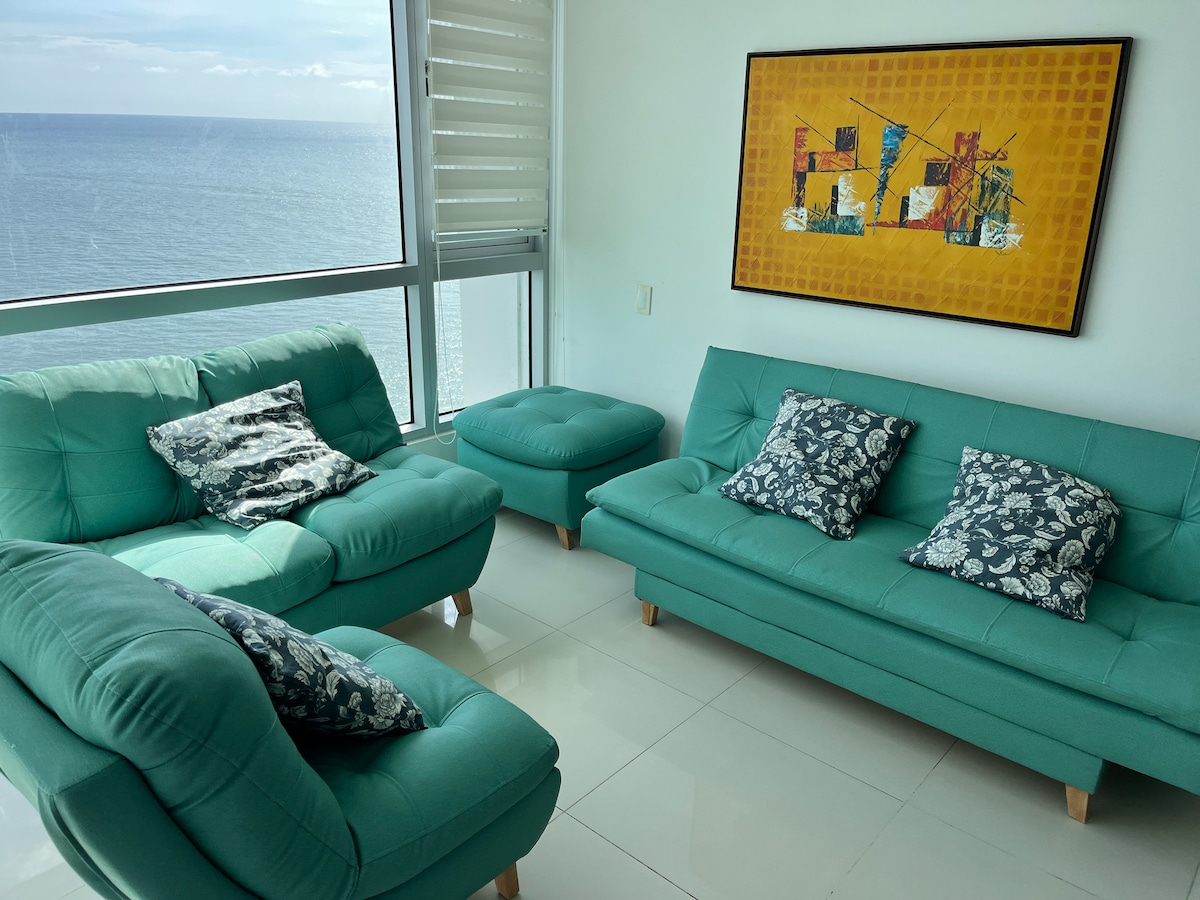 A bright living area features teal sofas and matching ottomans, complemented by decorative cushions. A large window offers views of the ocean, while a colorful piece of wall art adds a touch of creativity to the space.