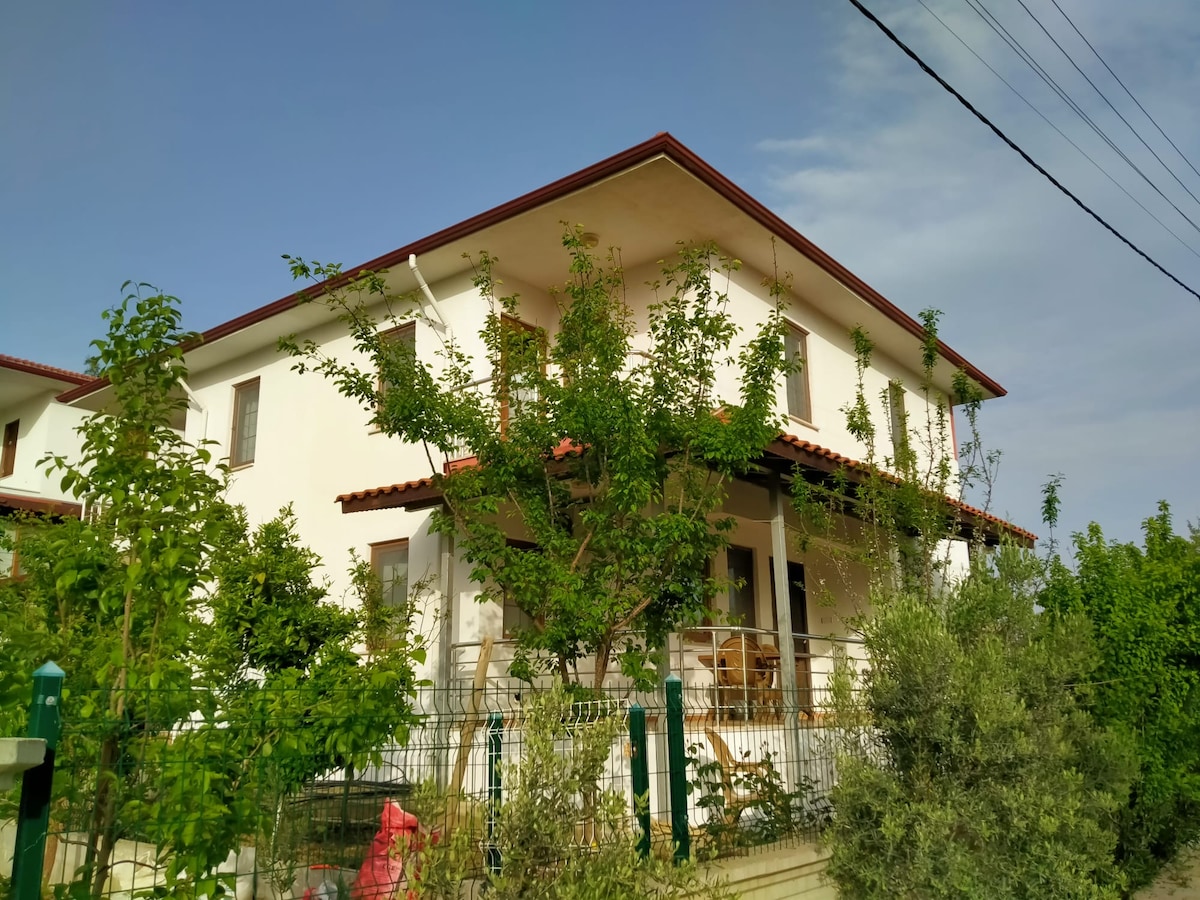 The two-story duplex home is set amidst lush greenery, featuring a welcoming porch shaded by overhanging trees. A white façade and red-tiled roof offer a charming contrast to the surrounding natural landscape, while a wooden railing frames the entrance.