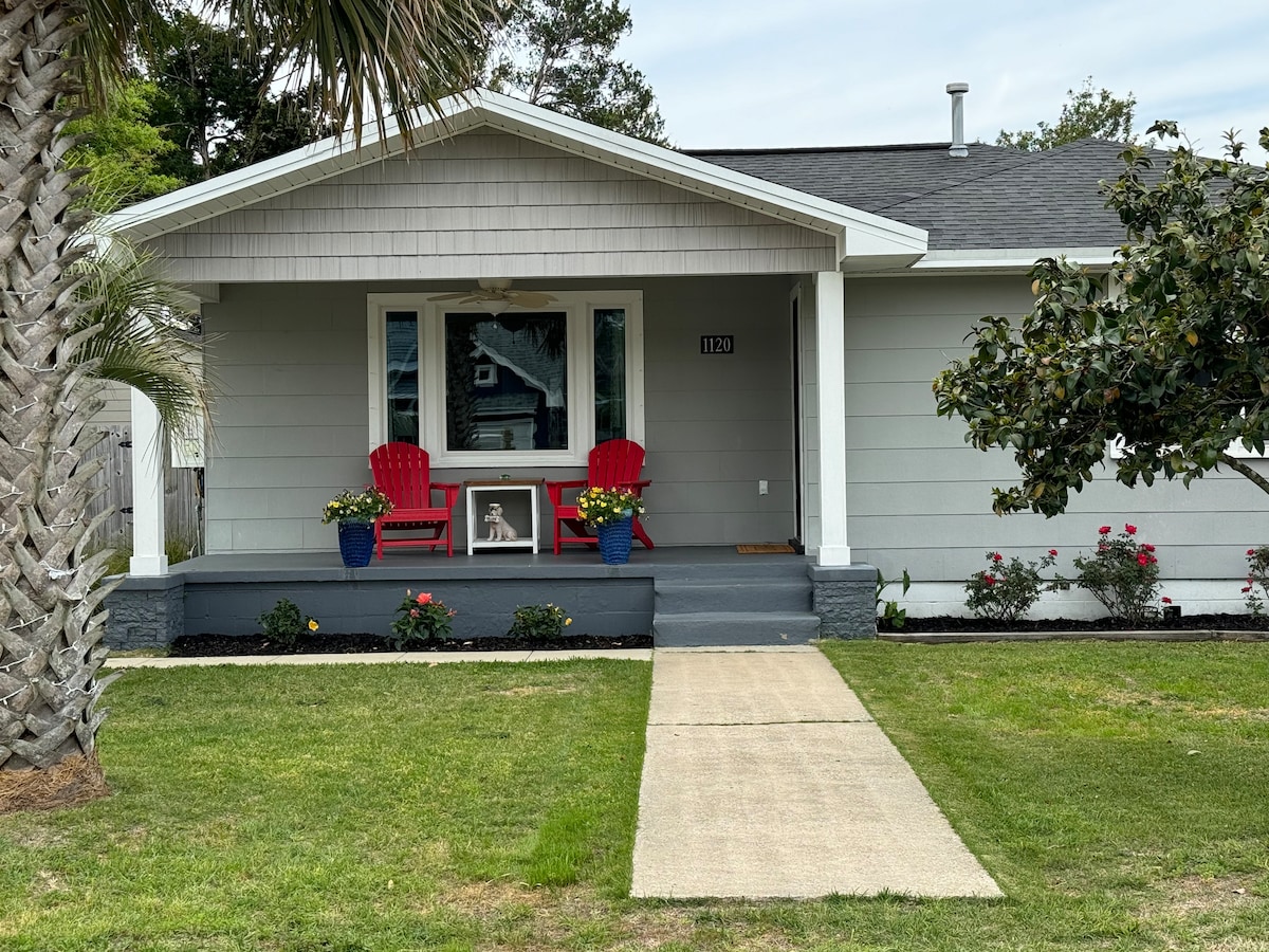 The exterior of the bungalow features a welcoming front porch with two red chairs flanking a small table. Potted plants with vibrant flowers are placed on either side of the entrance. A well-maintained lawn extends in front of the house against a light gray siding backdrop.