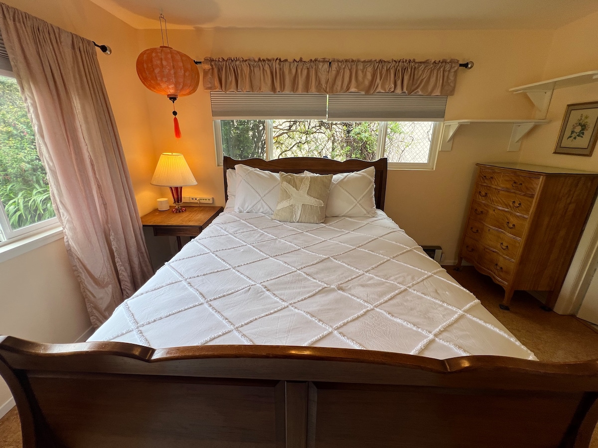 A cozy bedroom features a handmade wooden bed centered against the wall, dressed in a white quilt. Natural light filters through large windows adorned with light curtains, illuminating a bedside lamp and a wooden dresser. Soft decor elements create a soothing atmosphere for relaxation.