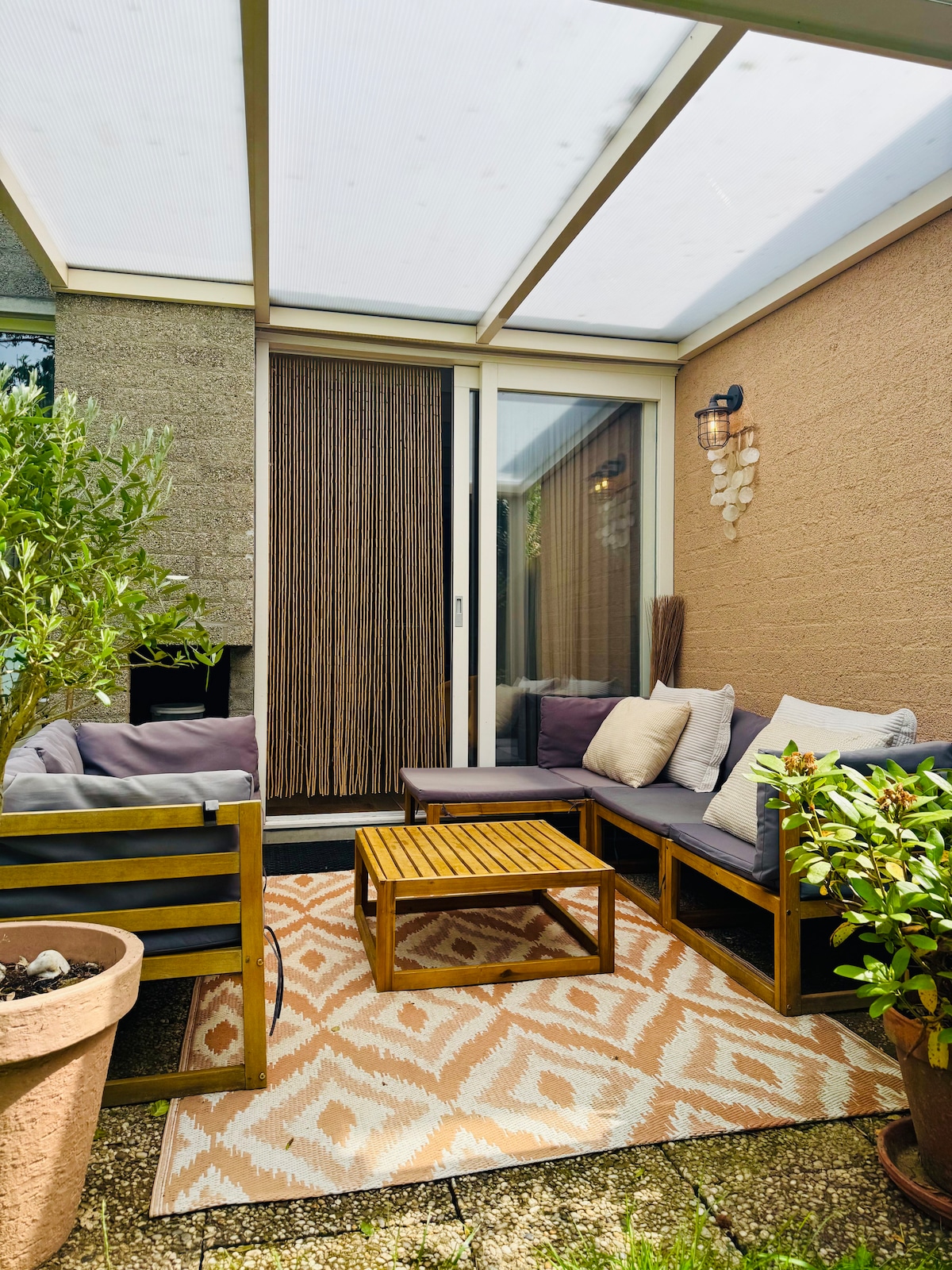 A sheltered outdoor seating area features a wooden coffee table surrounded by comfortable couches adorned with soft cushions. Decorative pots with greenery enhance the space, while a patterned rug adds warmth. The area is enclosed by a glass roof and walls, allowing for natural light.