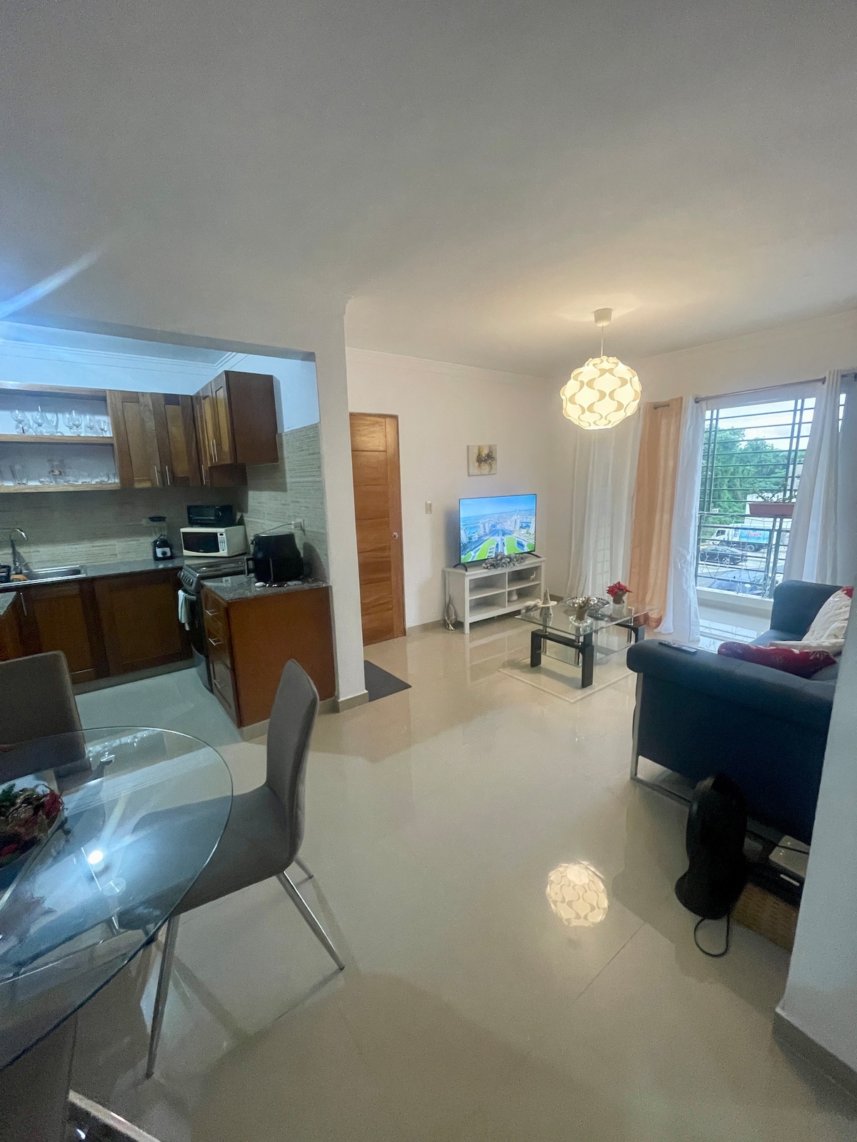 The image showcases a modern living area featuring a comfortable couch facing a television. A dining table with four chairs is positioned nearby. The kitchen is partially visible, equipped with wooden cabinets and appliances. Natural light flows in through large windows, enhancing the space.