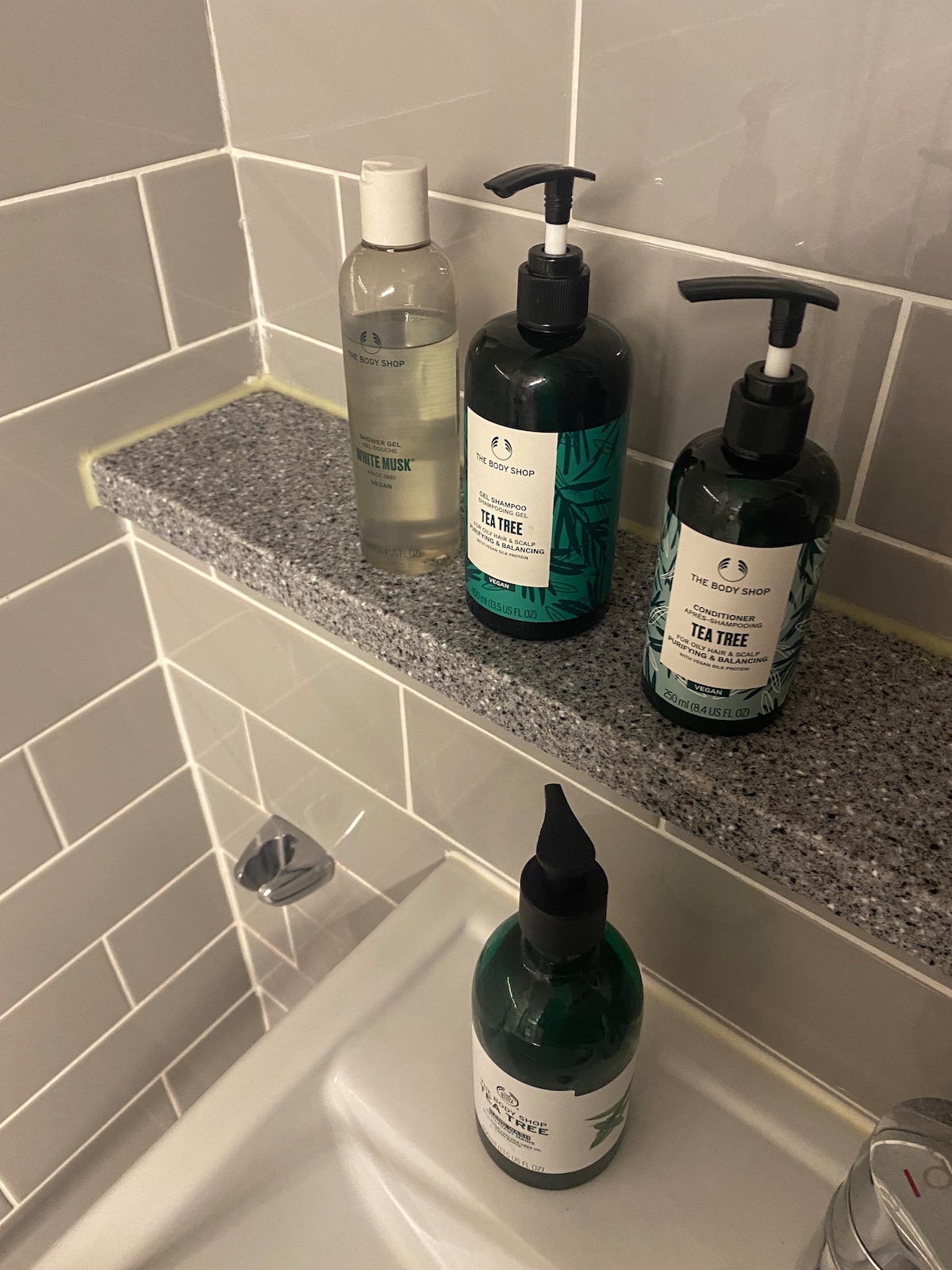 A shower shelf is visible, displaying four bottles of body care products. Two dark green bottles labeled 'Tea Tree' are positioned beside a clear bottle, while a black-topped bottle sits slightly to the right. The shelf is made of gray stone, set against white tiled walls.