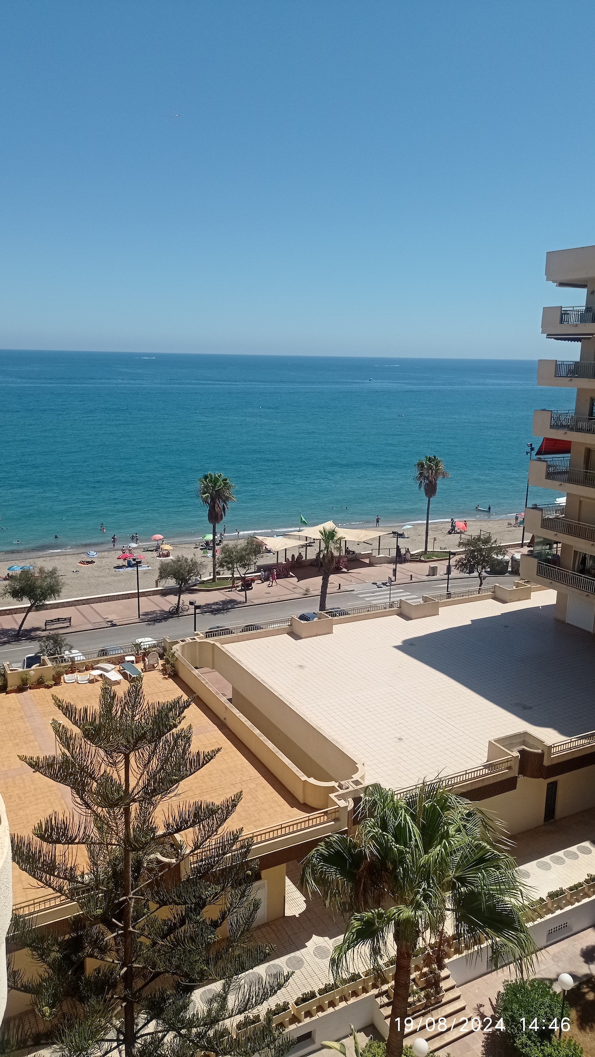 A view of the Mediterranean Sea is visible, with the coastline lined by palm trees and beachgoers. The sandy beach extends along the shore, while a clear blue sky complements the tranquil waters. A terrace area is seen below, enhancing the coastal ambiance.
