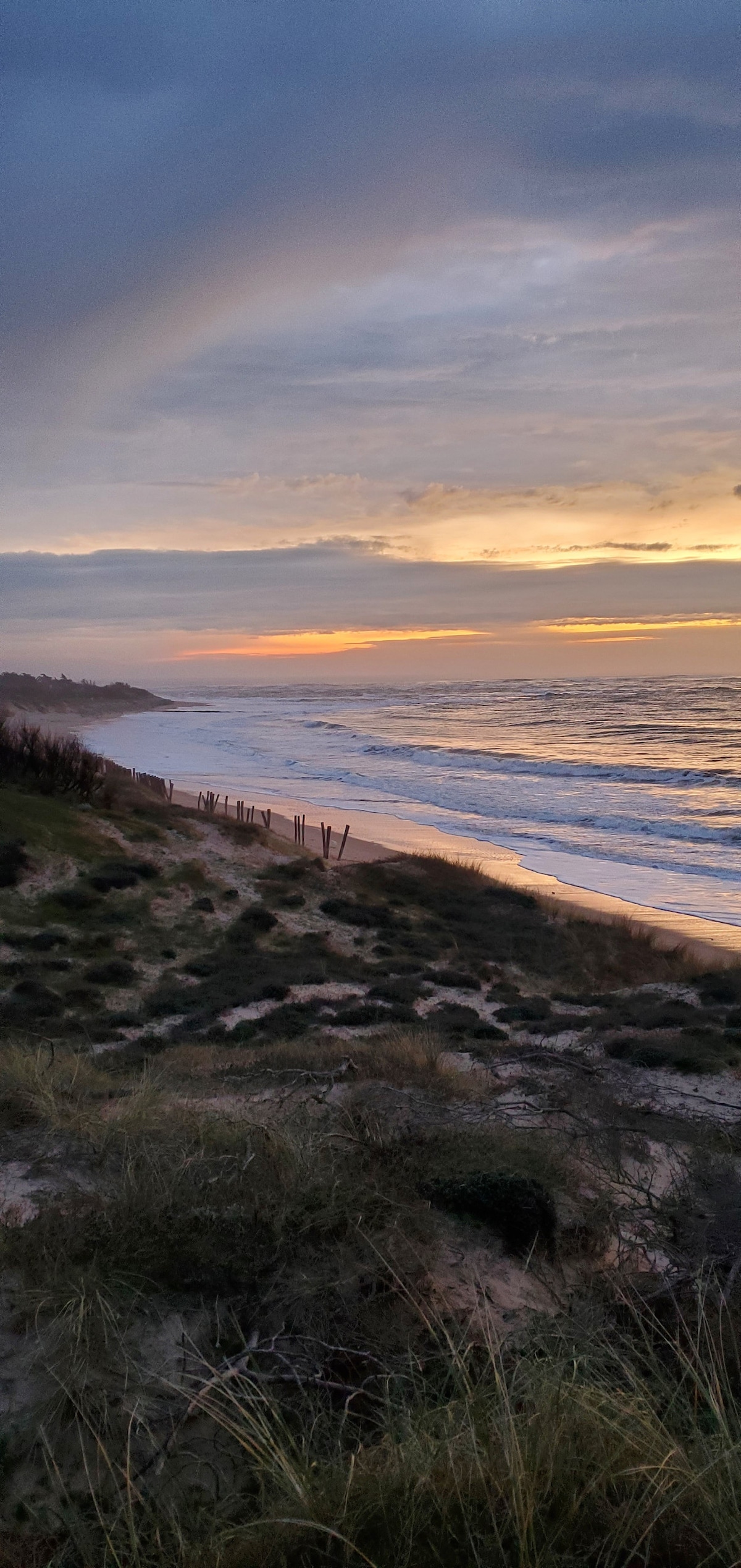 The shoreline is visible, framed by soft dunes and greenery. Gentle waves lap at the sand, reflecting the warm hues of sunset in the sky. Wispy clouds add texture to the scene, creating a serene coastal atmosphere.