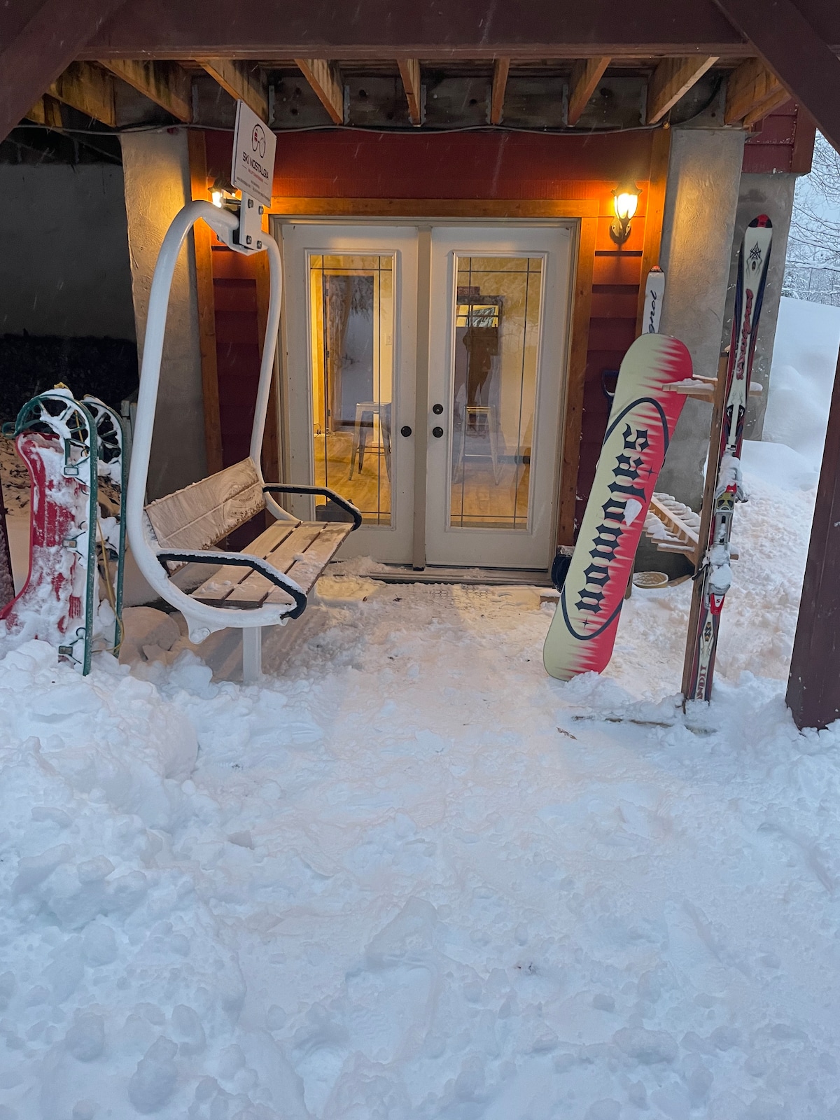 An entrance area is presented, flanked by snow and ski equipment. Two double doors, framed by warm lighting, lead into the chalet. A bench is positioned beneath the overhang, enhancing the welcoming ambiance of this ski-in/ski-out access point.