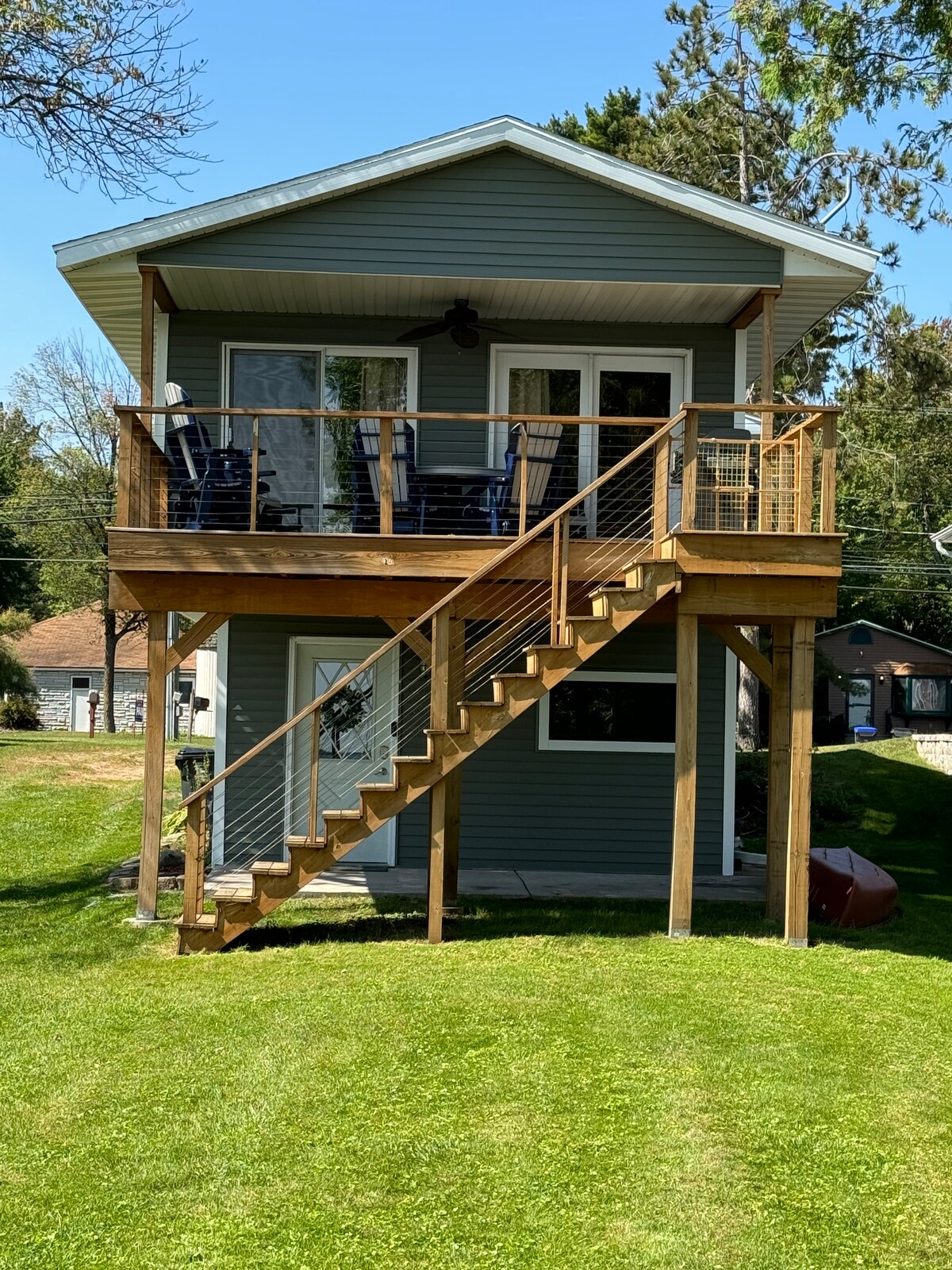 A two-story cottage is presented with a wooden deck and staircase leading to the doorway. The exterior features a soothing blue-grey color, complemented by large windows that allow natural light. The grassy area is well-maintained, providing a welcoming outdoor space.