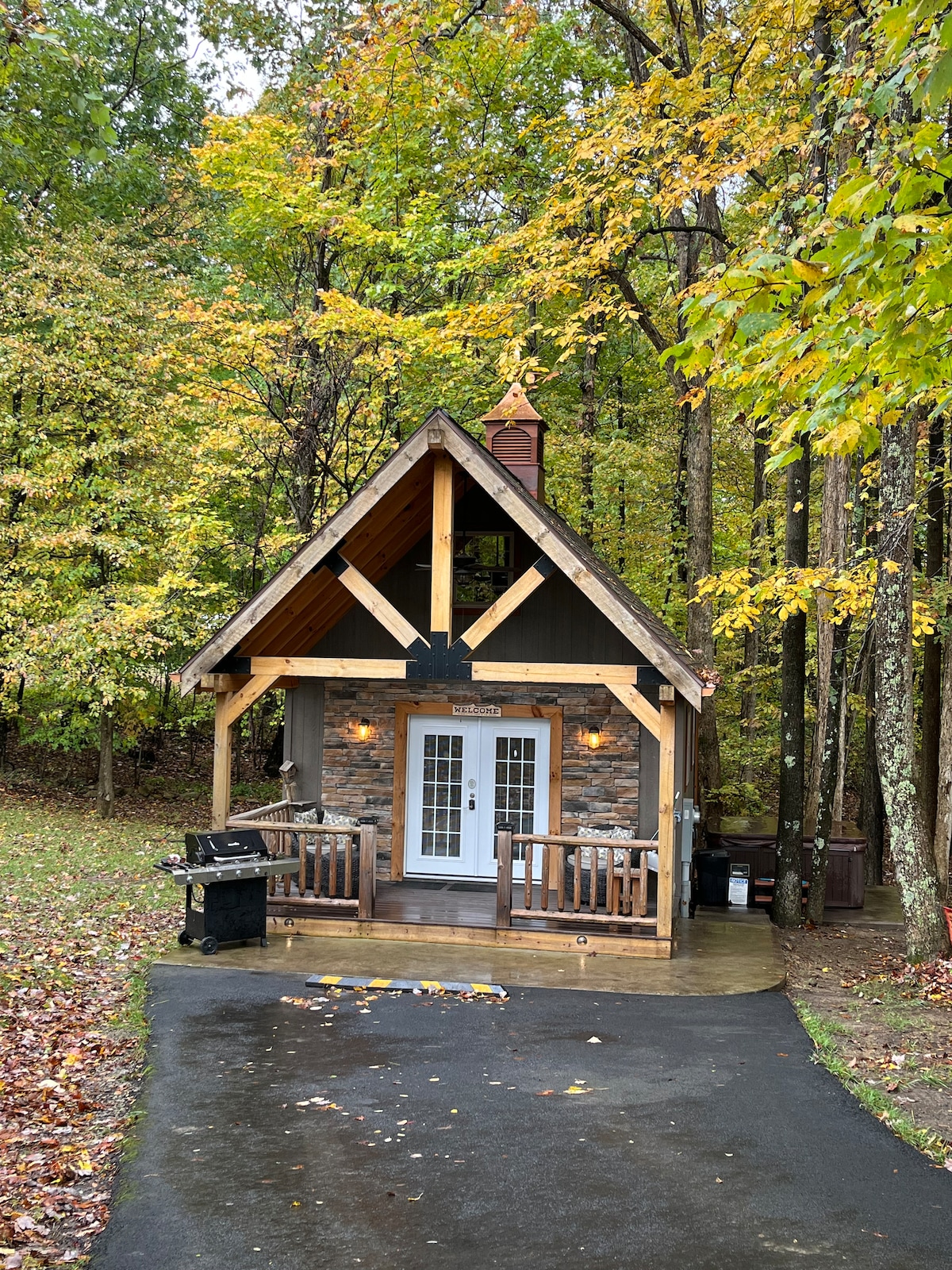 A charming cabin exterior is framed by vibrant fall foliage. The entrance features large glass doors flanked by warm lighting, while a wooden deck with a grill invites outdoor gatherings. The sloped roof is supported by wooden beams, contributing to the cozy aesthetic.
