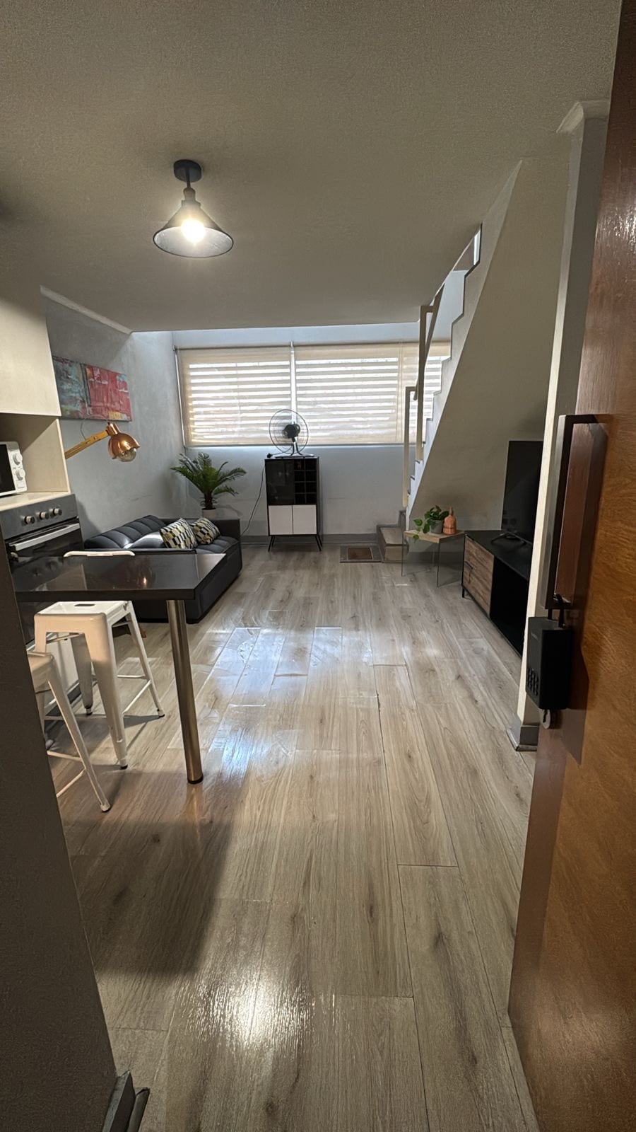 A view from the entrance reveals an open living space with a black couch and light wood flooring. Natural light enters through horizontal blinds. A staircase is visible on the right, while a small plant and decorative elements are placed throughout the area, adding to the inviting ambiance.