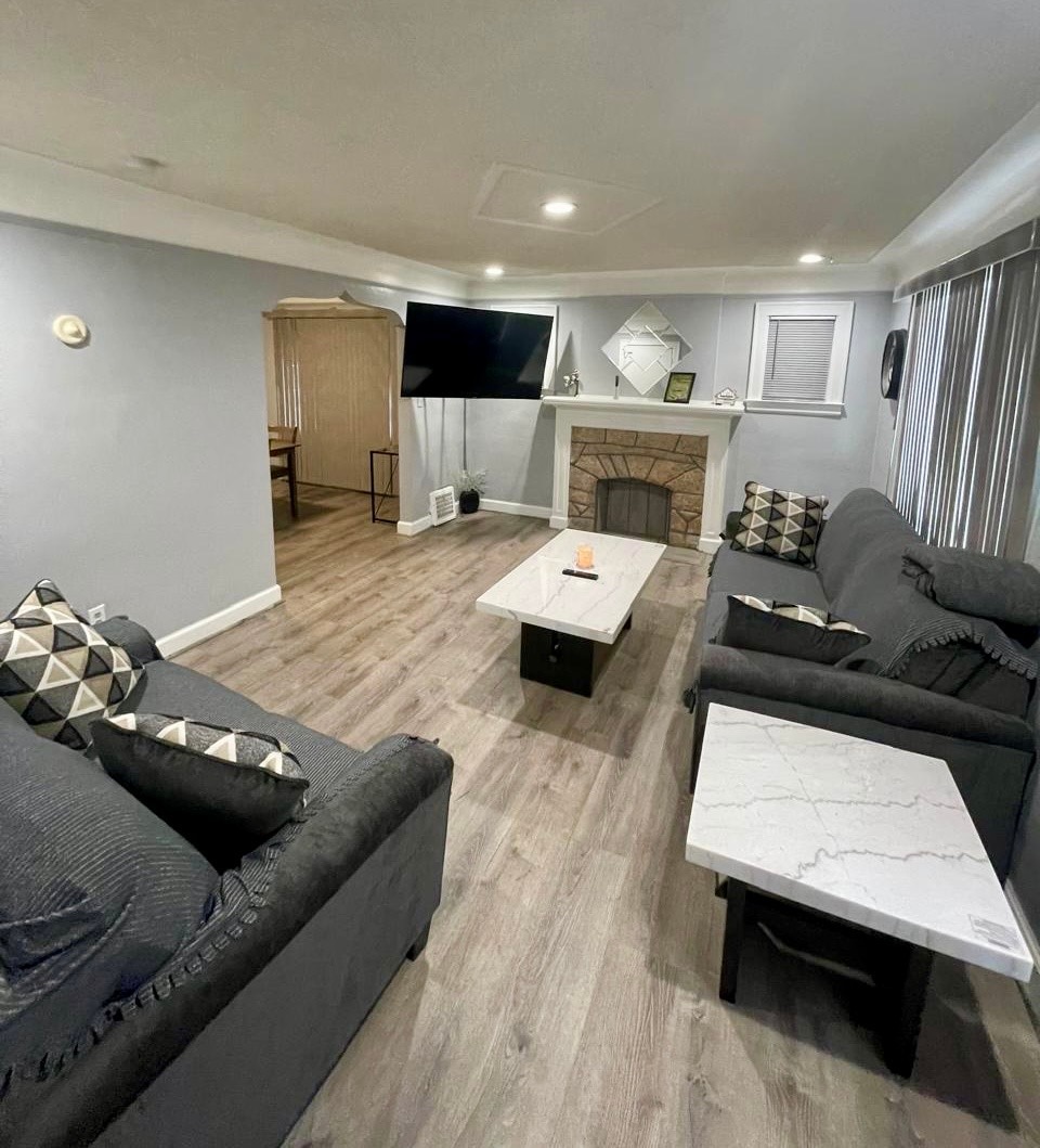 A spacious living area features two comfortable sofas adorned with patterned cushions. A coffee table with a sleek design sits between them, complementing the modern decor. A television is mounted above a decorative fireplace, with large windows allowing natural light to enter.