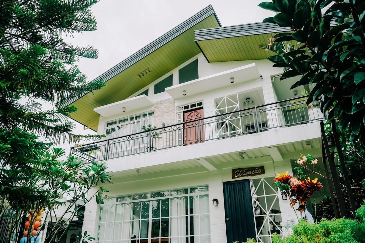 The exterior of a two-story house is shown, featuring a combination of modern architecture and lush greenery. Large windows allow for natural light, and a welcoming front door is highlighted. The balcony is accessible, offering a view of the landscaped surroundings.