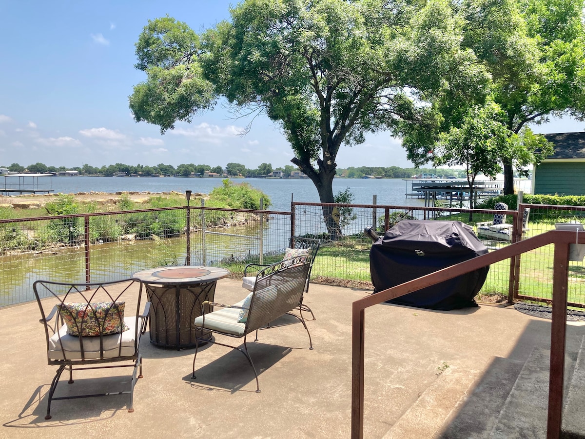 A private outdoor patio area features seating and a BBQ grill, surrounded by a fenced yard. This space overlooks the lake, framed by trees and greenery, providing a peaceful setting for relaxation and enjoying lake views.