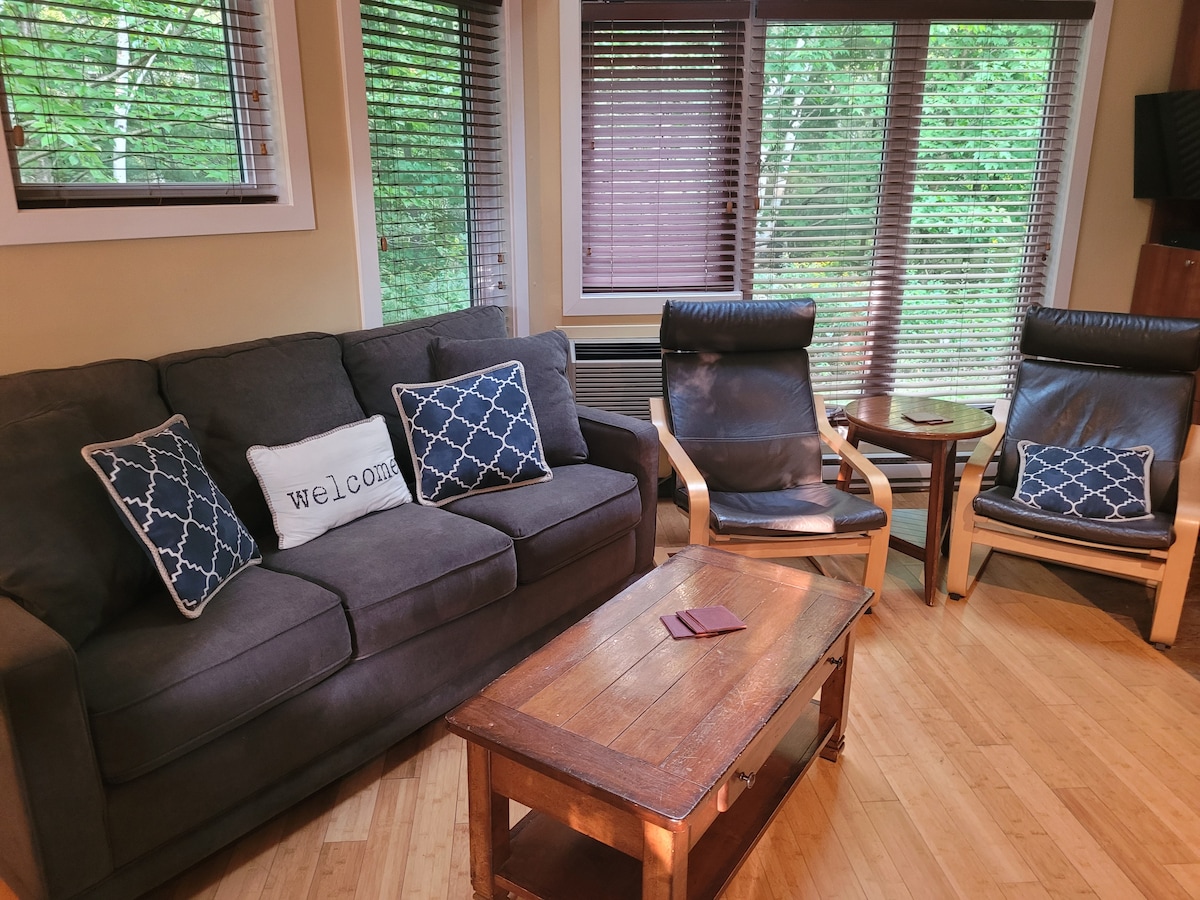 A comfortable seating area features a grey sofa with decorative pillows, paired with two armchairs and a wooden coffee table. Natural light filters through the window blinds, revealing a serene view of the surrounding greenery.