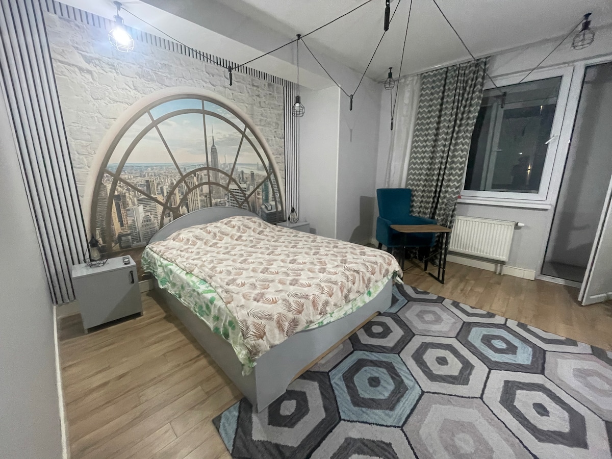 A spacious sleeping area features a large bed covered with a patterned blanket. A striking mural of a city skyline is displayed on one wall, while an accent chair and small table are positioned in the corner. Natural light streams through a window, with patterned curtains adding texture.