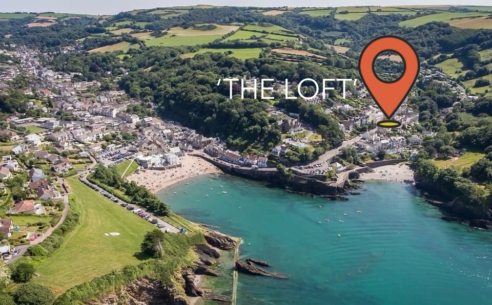 An aerial view captures the village of Combe Martin, surrounded by lush green hills, leading down to the sparkling blue sea. The coastline is dotted with sandy beaches and a lively village area, highlighting access to natural beauty and local amenities.