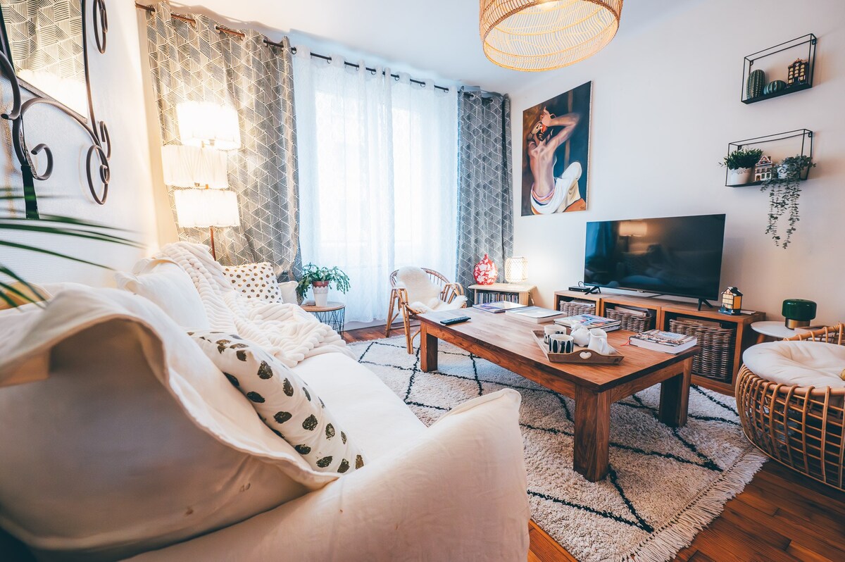 A living area is presented with a light, inviting sofa adorned with decorative pillows. A wooden coffee table sits centrally, complemented by greenery from potted plants. Soft lighting is provided by lamps, while a wall-mounted television offers entertainment options.