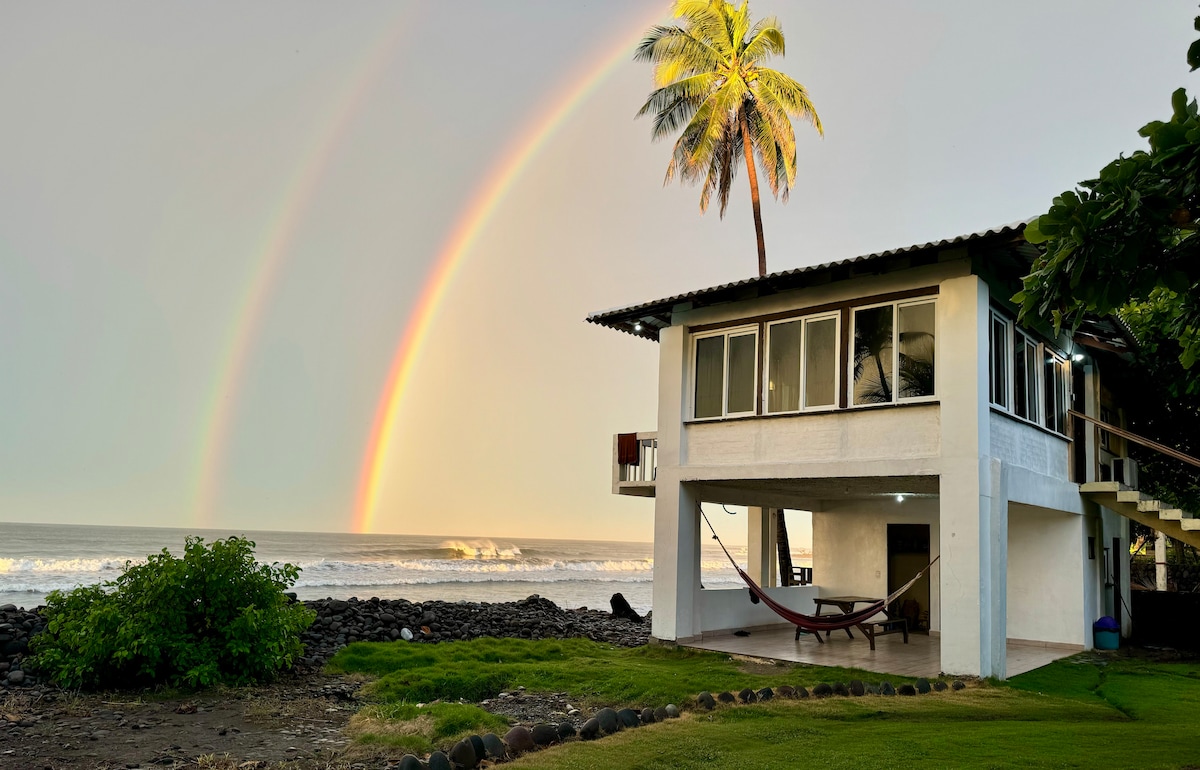 The house is positioned near the water, framed by palm trees and lush greenery. A vibrant rainbow arcs above, while the ocean waves crash in the distance. A hammock is visible under the spacious deck, providing a relaxing spot to enjoy the views.