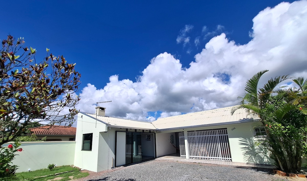 A single-story house is presented with a light-colored exterior and a gravel driveway. Lush greenery and blooming flowers surround the entrance. A clear blue sky with fluffy clouds enhances the welcoming appearance of the home.
