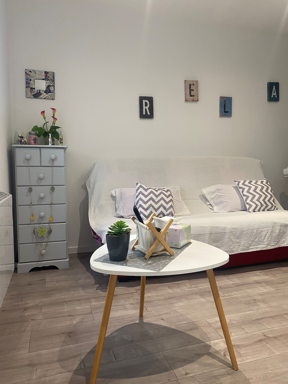 The living area contains a white sofa bed covered with a light throw. Decorative pillows in varied patterns are arranged on the sofa. A small round table with a plant centerpiece is positioned in front of the sofa. A light grey chest of drawers stands against the wall, adorned with simple decorations.