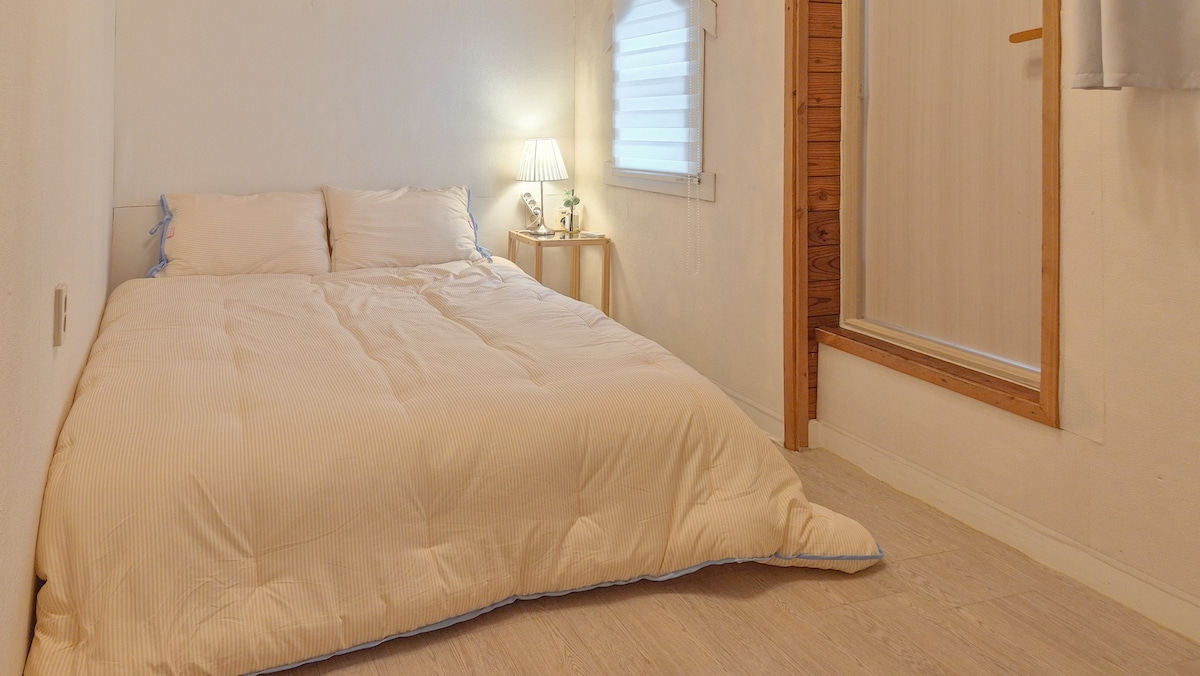 A well-lit sleeping area features a spacious bed with a soft, light-colored duvet. A small bedside table with a lamp is present beside the bed. A wooden door leading to the bathroom is positioned on the right.