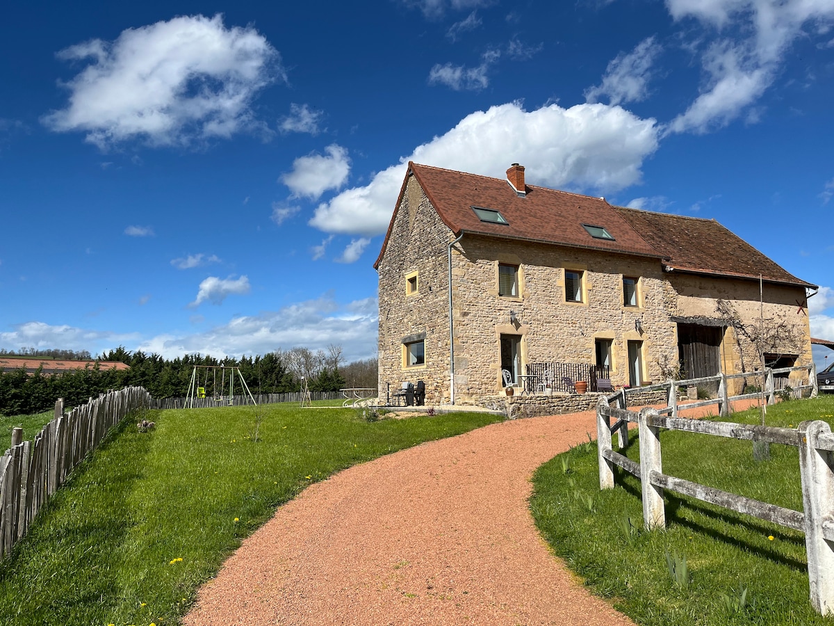 A rustic farmhouse is set against a bright blue sky, showcasing its stone façade and red roof. A gravel path leads to the entrance, bordered by green grass and wooden fencing. Lush fields and trees are visible in the background, creating a serene countryside setting.