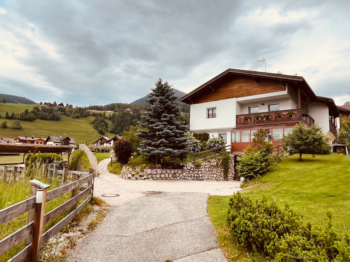 A charming two-story building is situated among lush greenery, featuring a wooden balcony adorned with potted plants. A pathway lined with stone walls leads to the entrance, with rolling hills and trees visible in the background under an overcast sky.