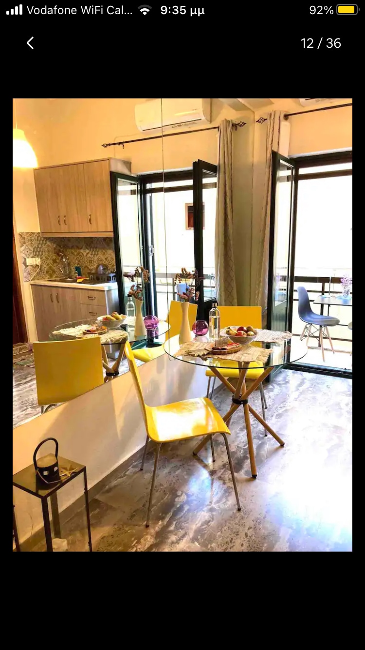 A modern dining area is presented with a glass table and four yellow chairs. A bouquet of flowers adds a touch of color. Large glass doors open to a small balcony, allowing natural light to fill the space. The kitchen area is visible in the background, featuring wooden cabinetry.