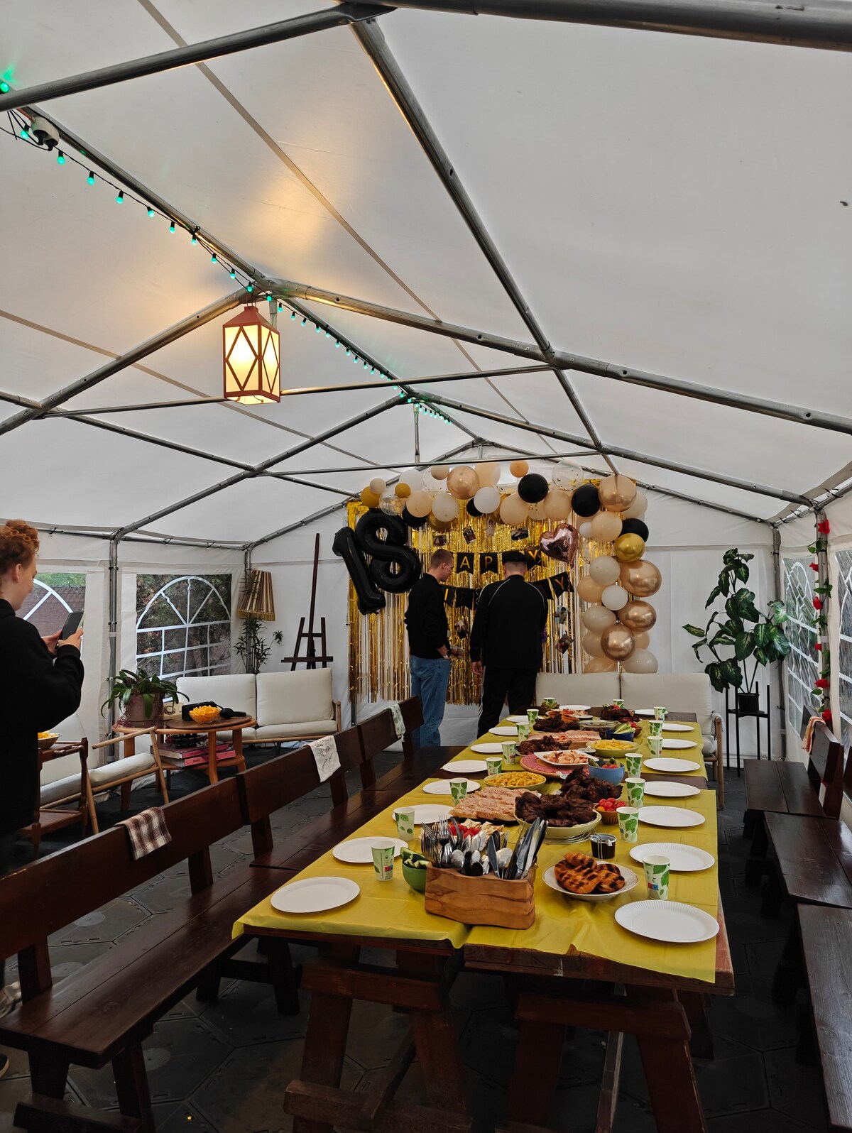 An expansive party tent is set for a gathering, featuring a long banquet table adorned with brightly colored plates and an array of food. Decorative balloons and a photo display enhance the festive atmosphere, complemented by wooden benches around the table.