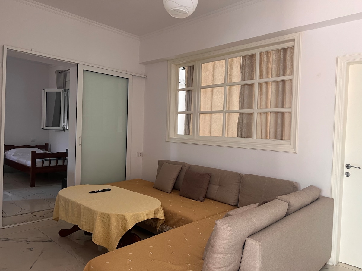 A living area is displayed, featuring a cozy sectional sofa with neutral tones and several cushions. A dining table covered with a light fabric sits nearby, complemented by a remote control. Large windows allow natural light to fill the space, connecting to a visible bedroom area.