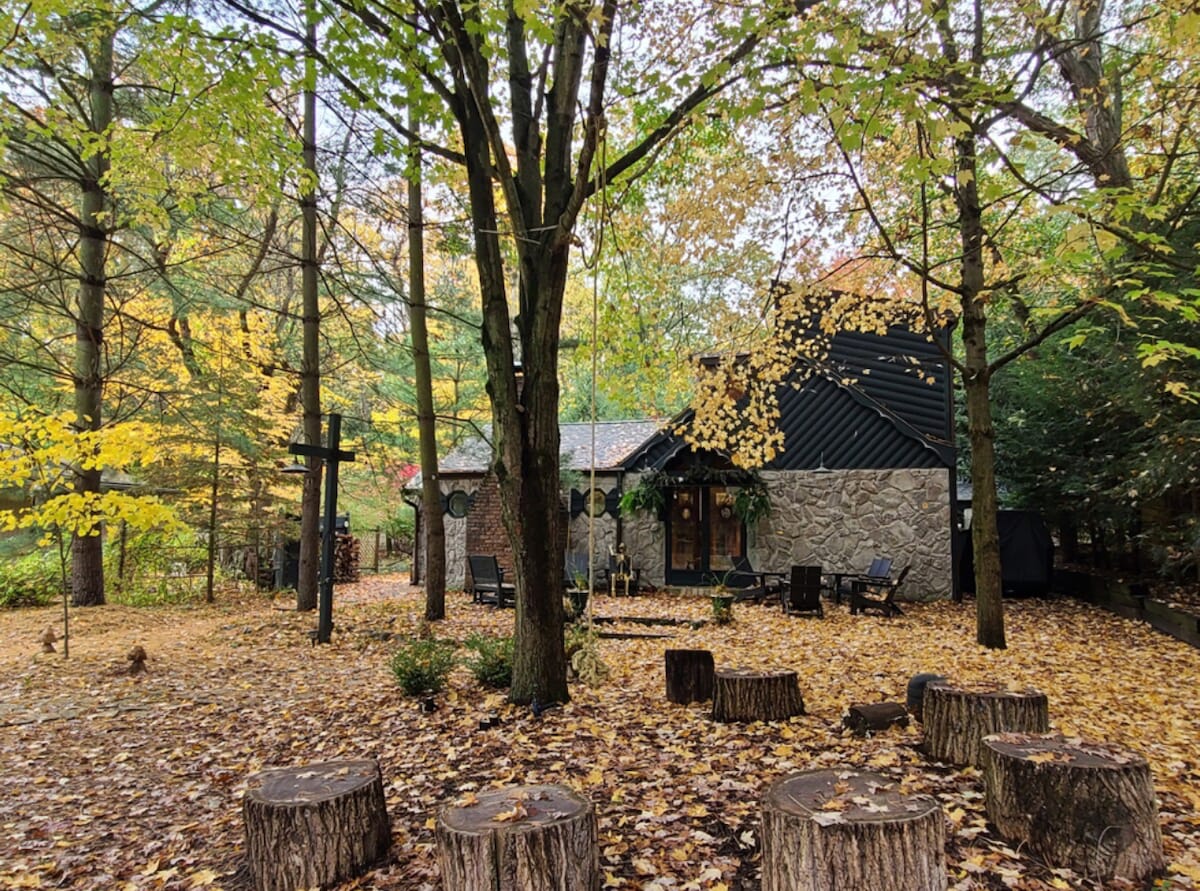 A charming cottage is surrounded by a vibrant autumn landscape, with golden and orange leaves contrasting against a dark structure. The yard features a circle of wooden stumps and a few seating areas, giving a cozy feel amidst the natural setting.