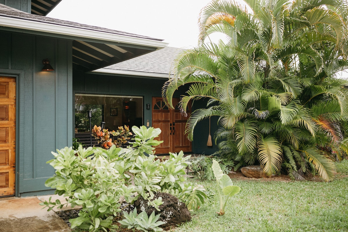The exterior of the home features a vibrant garden area with tropical plants and lush greenery. A wooden door with intricate designs provides access to the inviting entrance. The roof overhang offers a sheltered area, enhancing the home's tropical aesthetic.