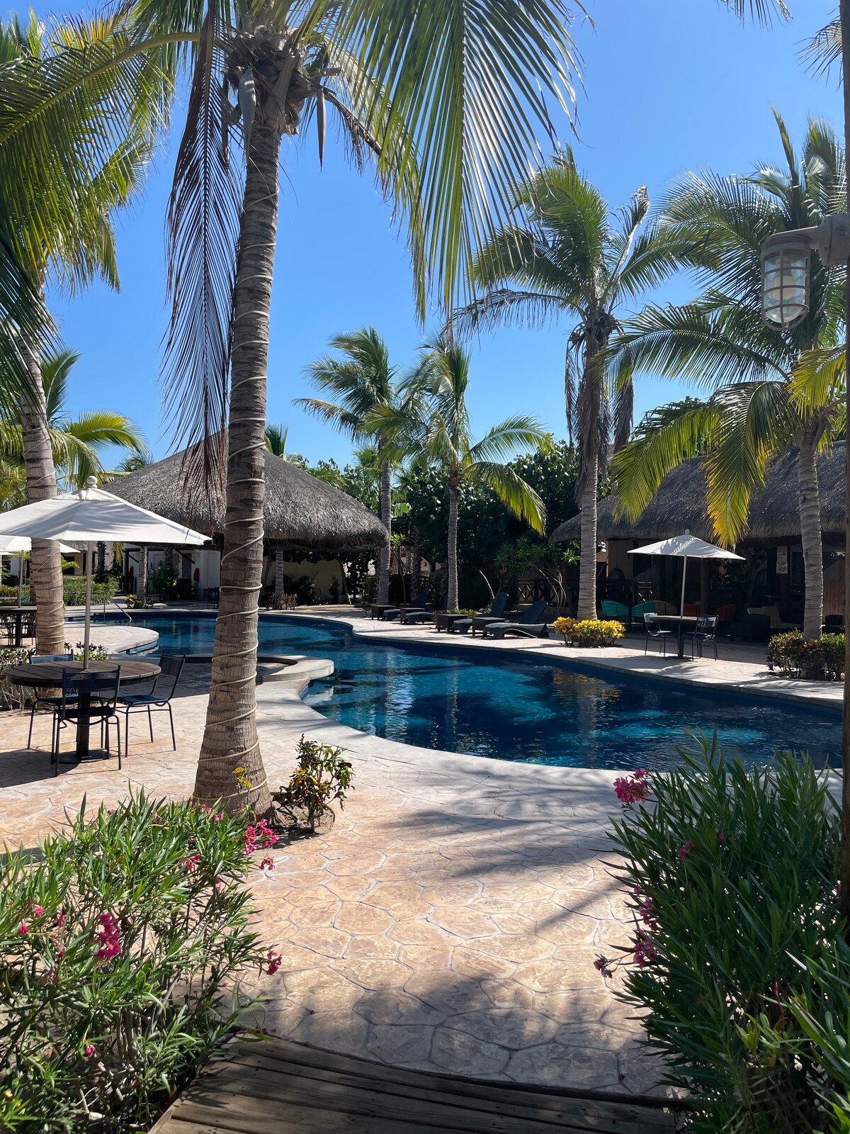 Surrounding palm trees provide shade near a large swimming pool, which reflects the clear blue sky. Various outdoor seating areas are visible, featuring tables and umbrellas. Lush greenery and flowering plants add color to the landscape, enhancing the serene atmosphere.