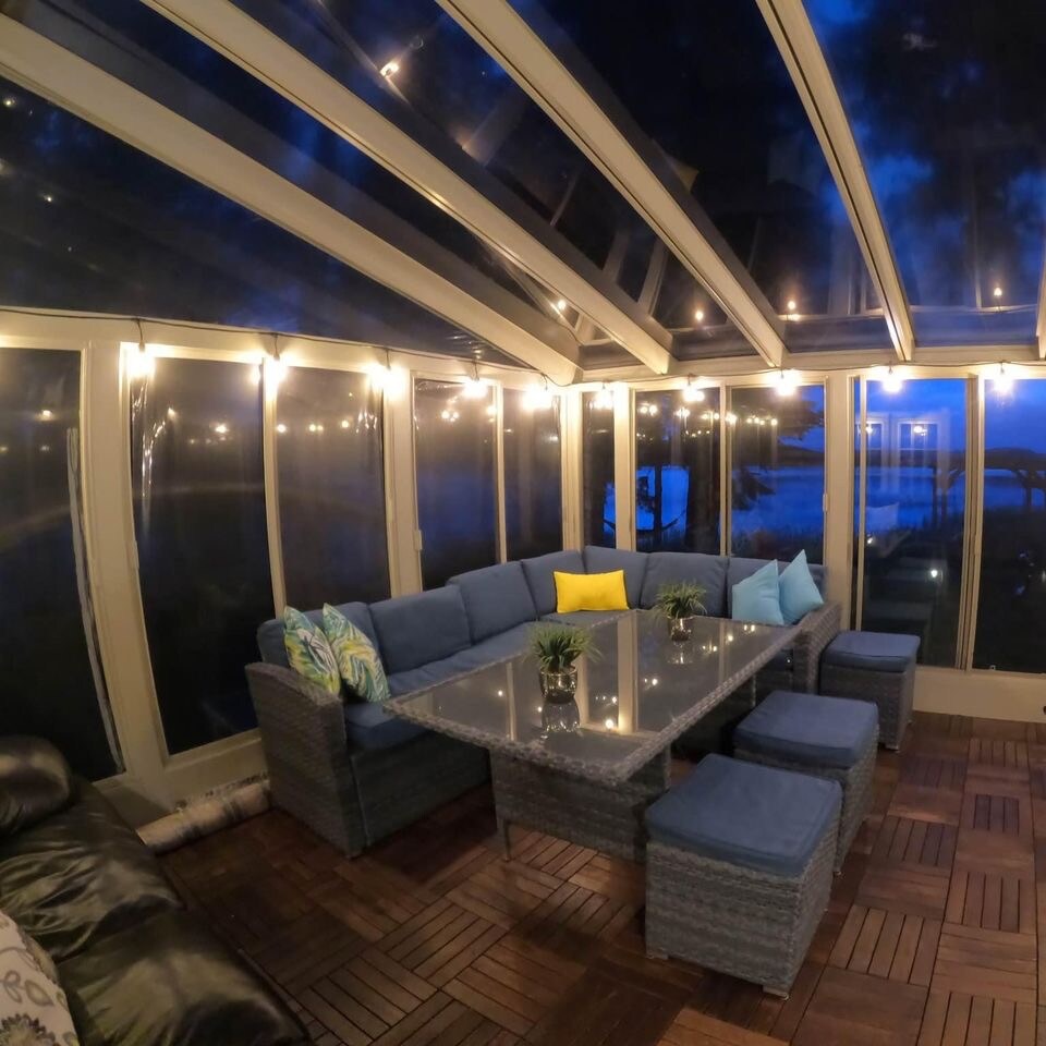 A spacious solarium features a large glass table surrounded by comfortable seating, enhanced by soft lighting. The floor is made of wooden tiles, and plants are placed decoratively on the table. Views of the surrounding nature are visible through the glass walls.