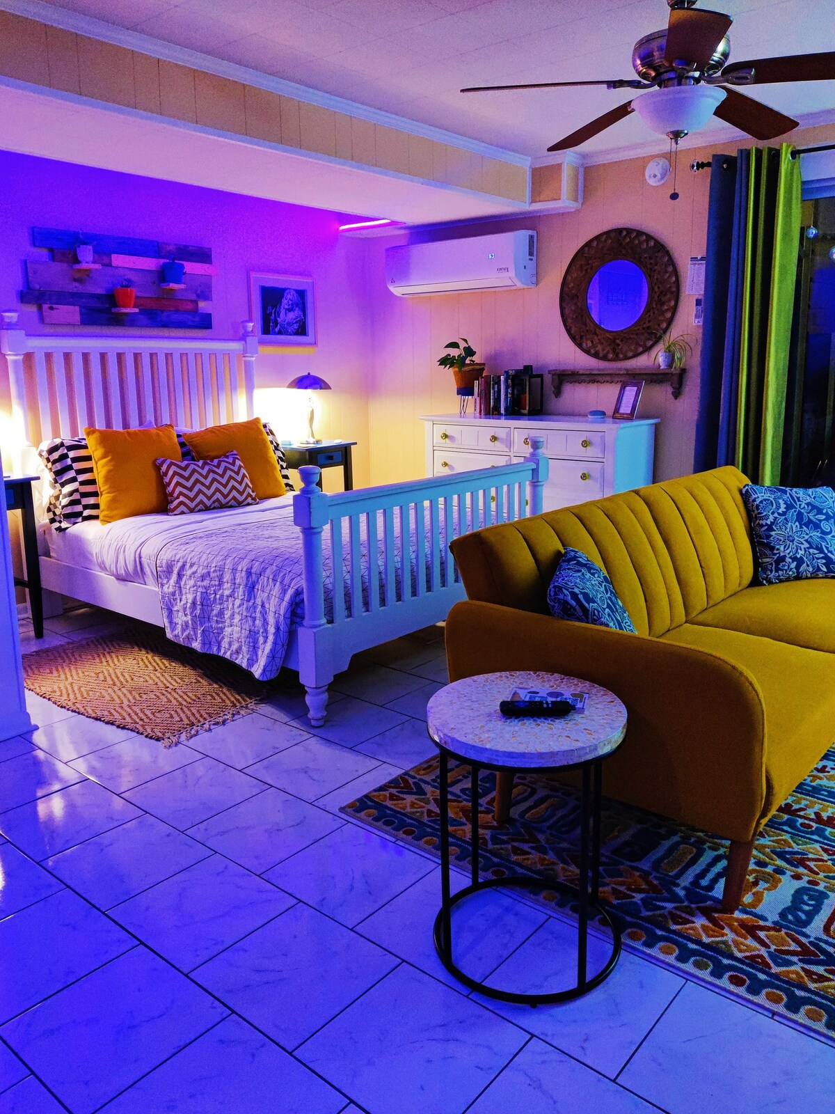 A cozy studio space features a white bed adorned with layered blankets and pillows. A vibrant yellow sofa and patterned area rug add color. Soft lighting creates a warm ambiance, complemented by decorative accents and a dresser with neatly arranged items.