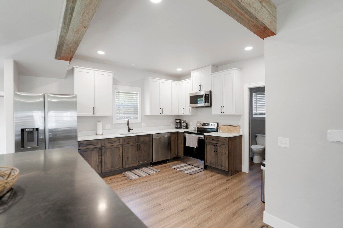 A modern kitchen is highlighted with white cabinetry and stainless-steel appliances. A sleek countertop offers ample space, complemented by wood accents. Under-cabinet lighting enhances the workspace, while a window provides natural light. Light wood flooring adds warmth to the open design.