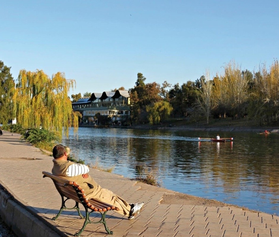 A peaceful riverside scene features a person relaxing on a bench under the shade of trees. The calm water is home to rowers gliding by, with a building visible in the background, surrounded by greenery.