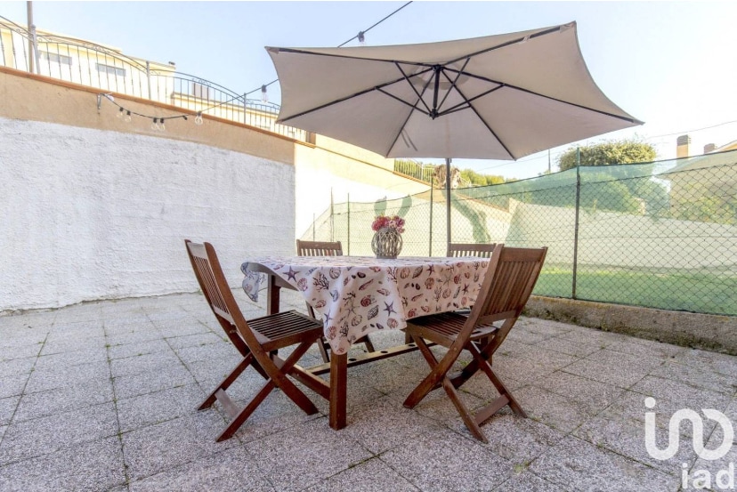 An outdoor dining area is featured, consisting of a wooden table surrounded by four folding chairs. A large umbrella provides shade overhead, while a small vase with flowers adds a decorative touch. A grassy area is visible in the background, enclosed by a fence.