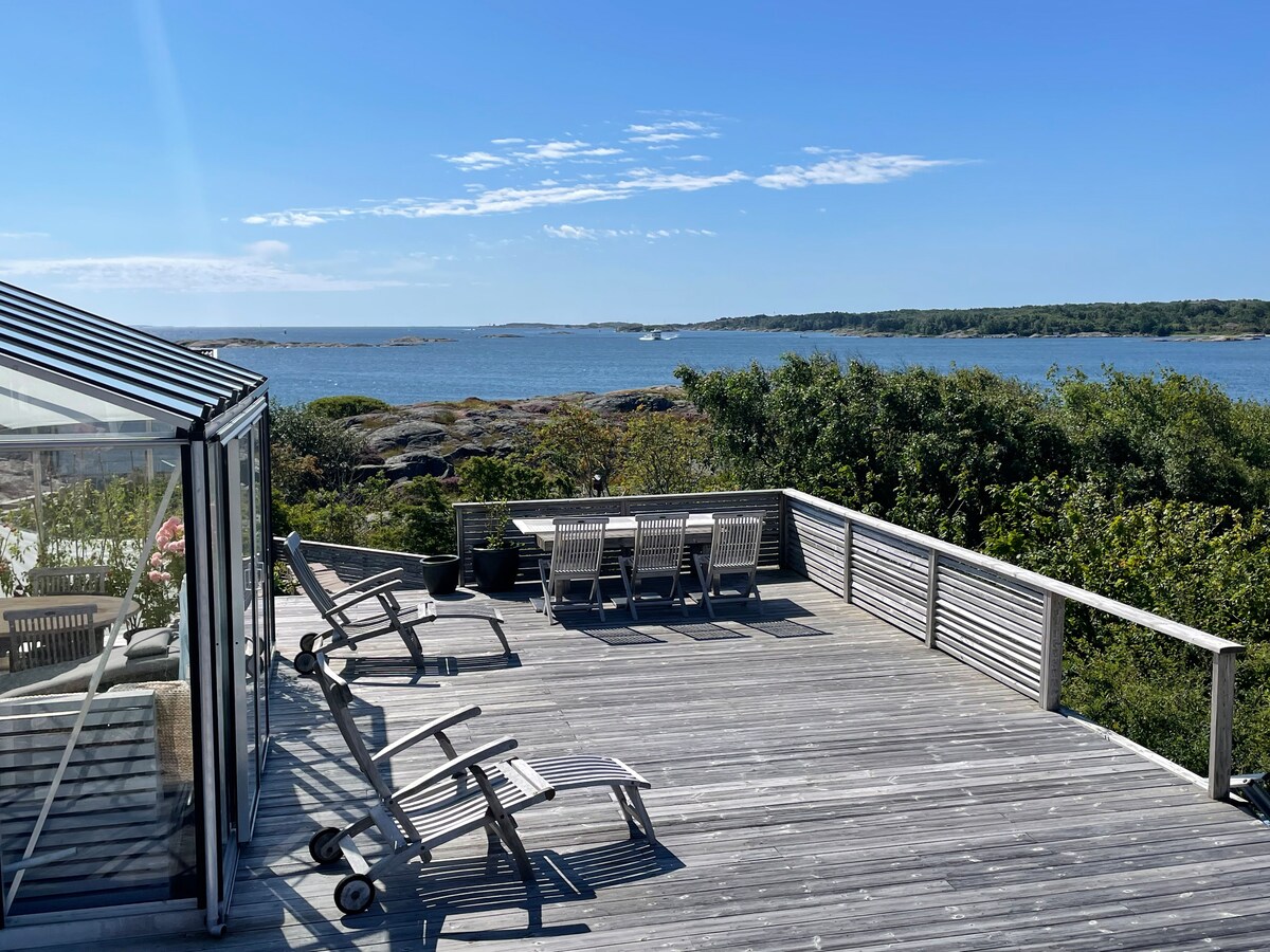 A spacious deck offers panoramic views of the surrounding landscape and coastline. Several lounge chairs are positioned for relaxation, while a dining table accommodates outdoor meals. The scene includes greenery and rocky formations, enhancing the natural setting by the water.