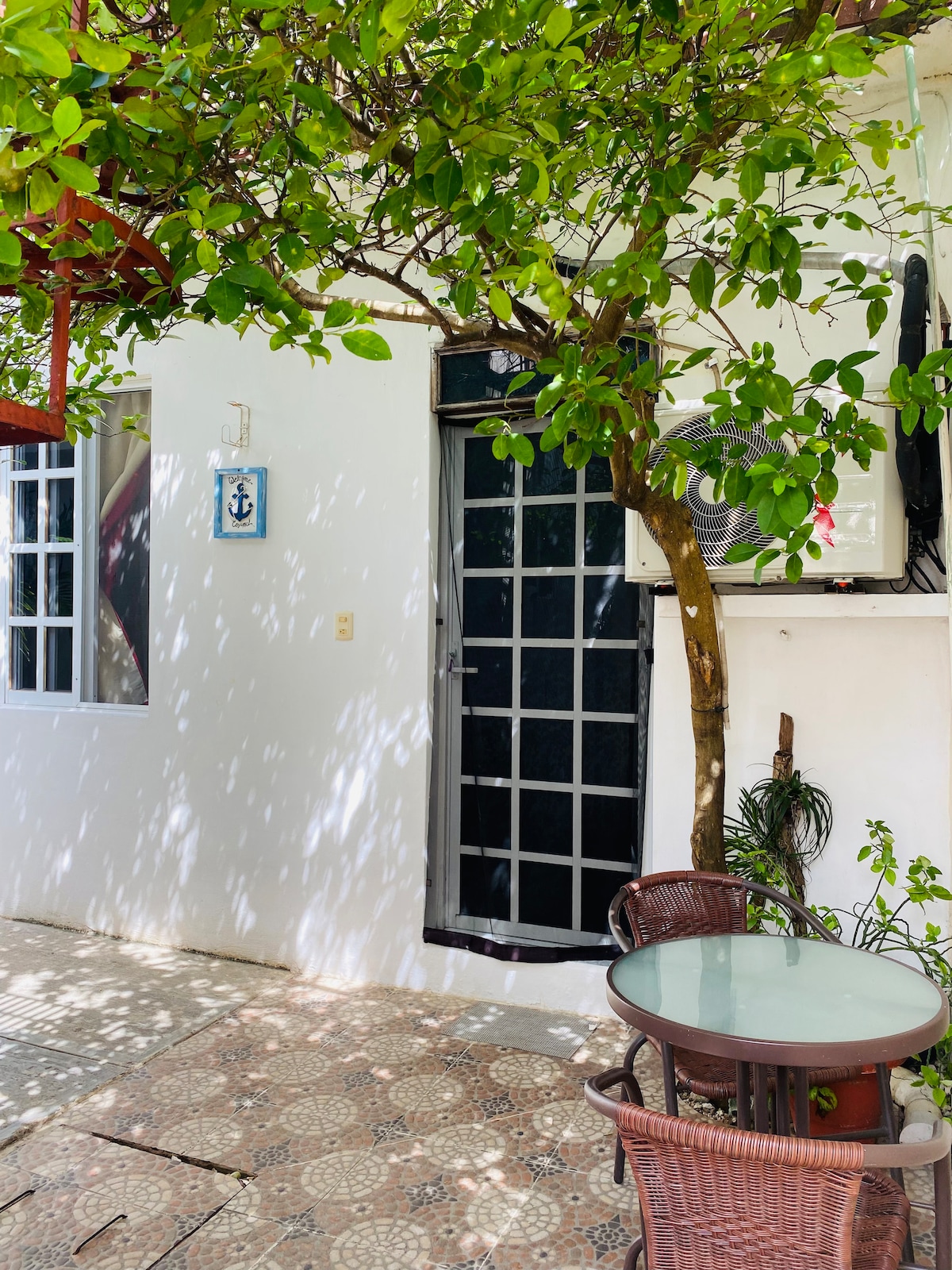 A shaded outdoor area features a small round table with two woven chairs, surrounded by dappled sunlight filtering through tree leaves. The entrance is highlighted by a black door, flanked by white walls and a decorative blue anchor wall hanging.