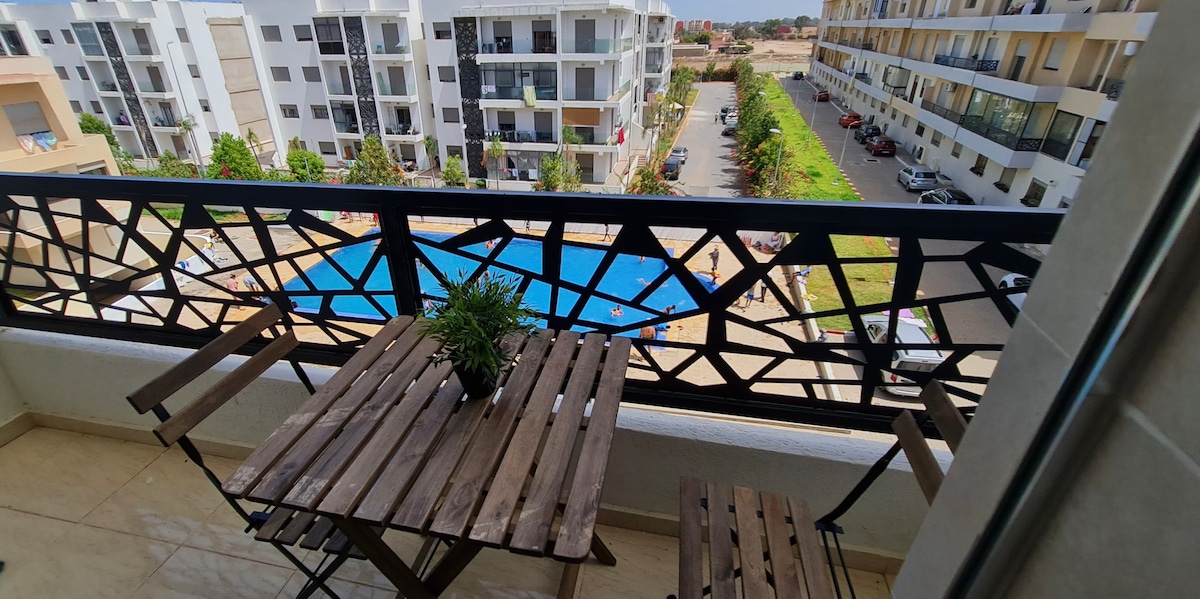 A balcony offers a view of a shared pool surrounded by landscaped areas. Two wooden chairs and a small table are placed unobtrusively, alongside a potted plant, enhancing the outdoor seating experience.