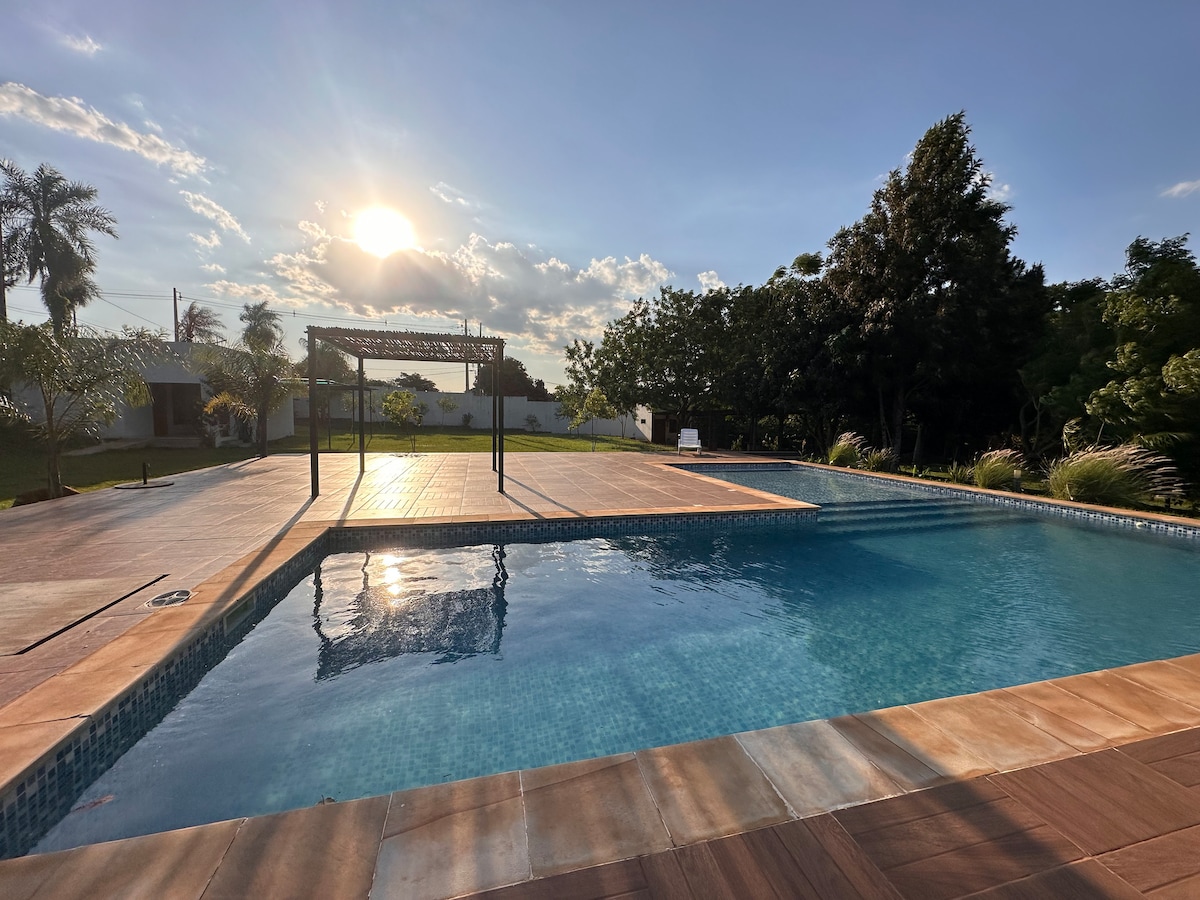 A spacious outdoor pool area is visible under a clear sky, with sunlight reflecting on the water's surface. Surrounding greenery and a shaded seating structure enhance the tranquil setting, making it suitable for relaxation.