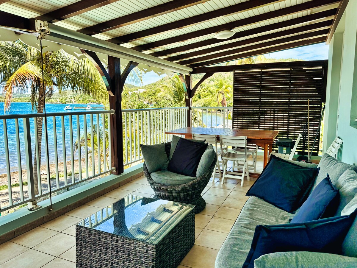 A spacious outdoor terrace is presented with a comfortable seating area, featuring a large table and cozy chairs. Views of the calm ocean and palm trees are visible through the railing. The shaded area offers a relaxing spot to enjoy the surroundings.