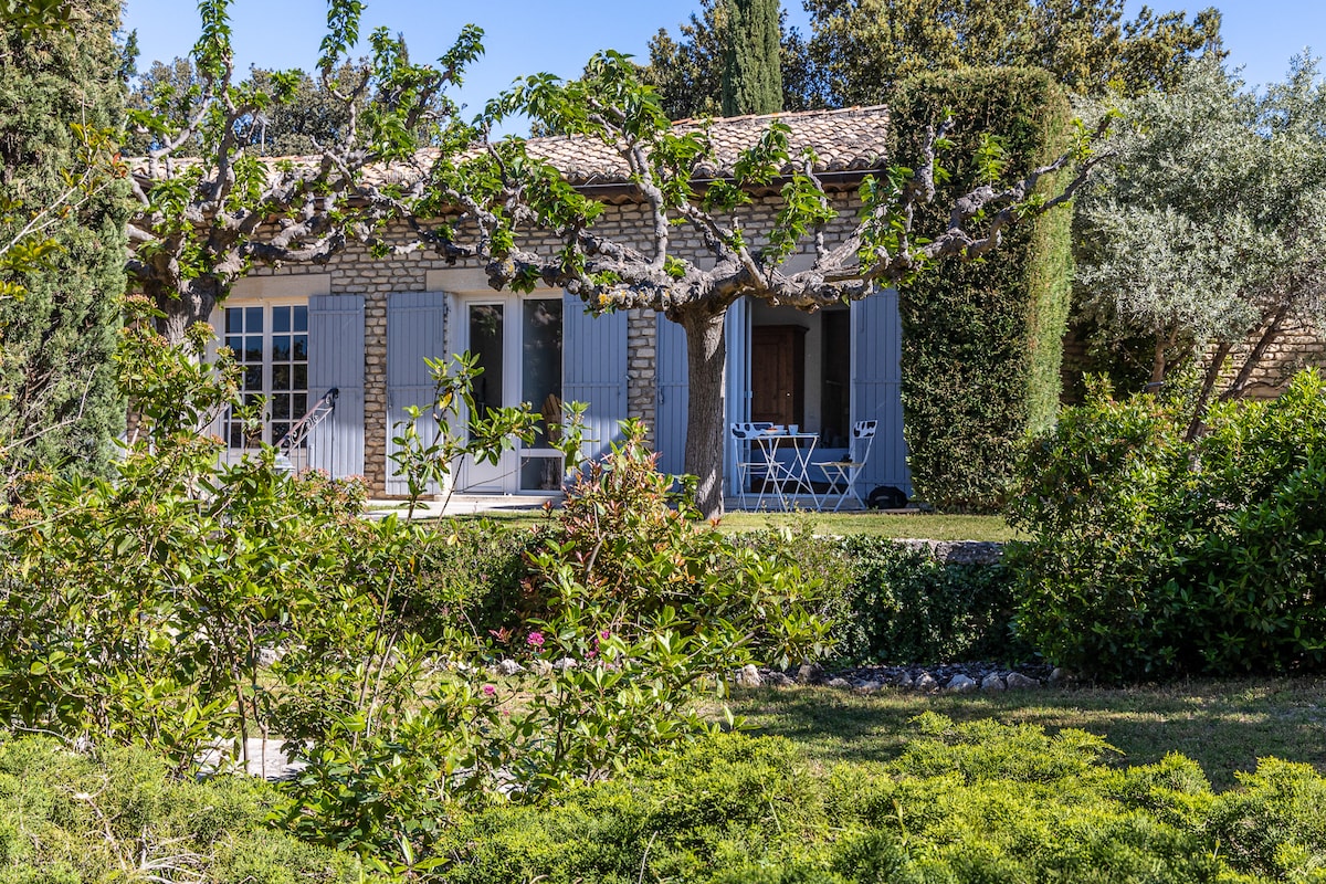The stone house is set amidst lush greenery, featuring light blue shutters and a tiled roof. A small outdoor seating area with a table and chairs is visible on the terrace, framed by flowering plants and trees. A sunny ambiance characterizes the exterior.